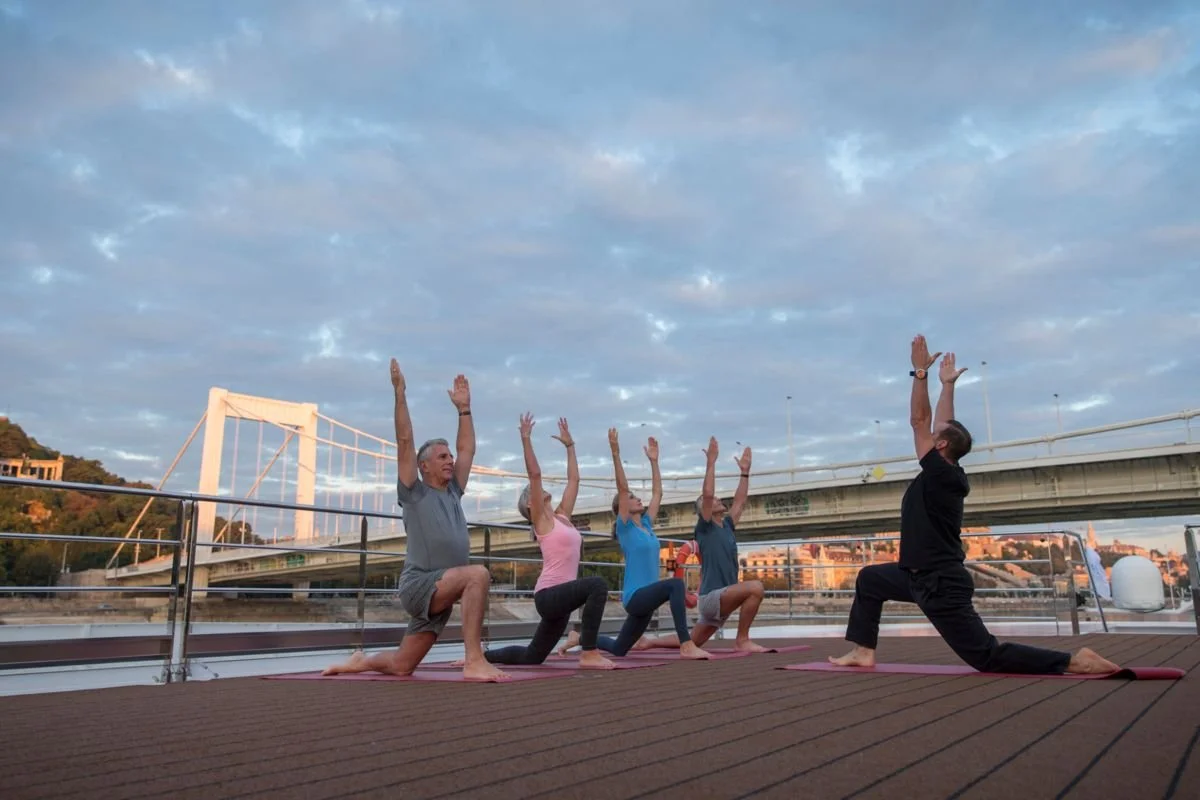 Group of five people practicing yoga on a deck near water with a city in the background, during sunset.