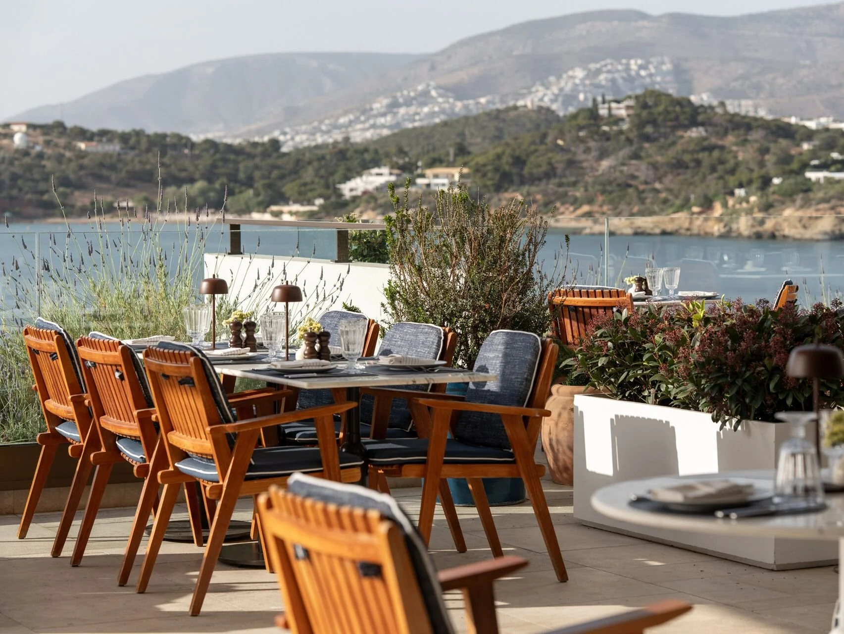 Outdoor restaurant patio with wooden chairs and table set with glassware and tableware, overlooking a body of water and hills in the background.