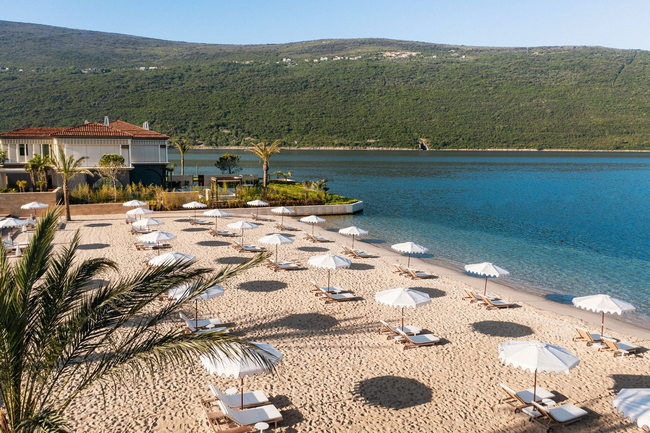 A scenic view of a resort beach with white umbrellas and lounge chairs on sandy shore next to a calm water body, green hills in the background, and a modern building with a red-tiled roof.