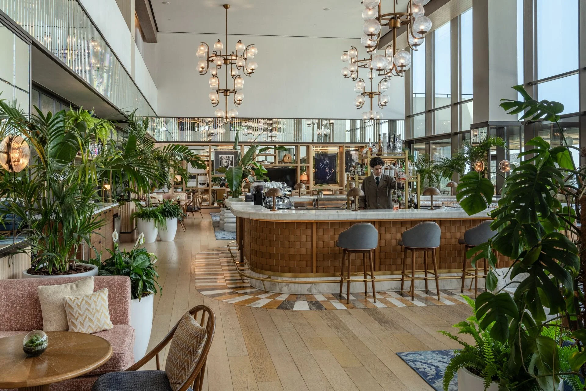 Interior of a modern, stylish cafe with large plants, wooden furniture, a marble counter, and contemporary chandeliers.