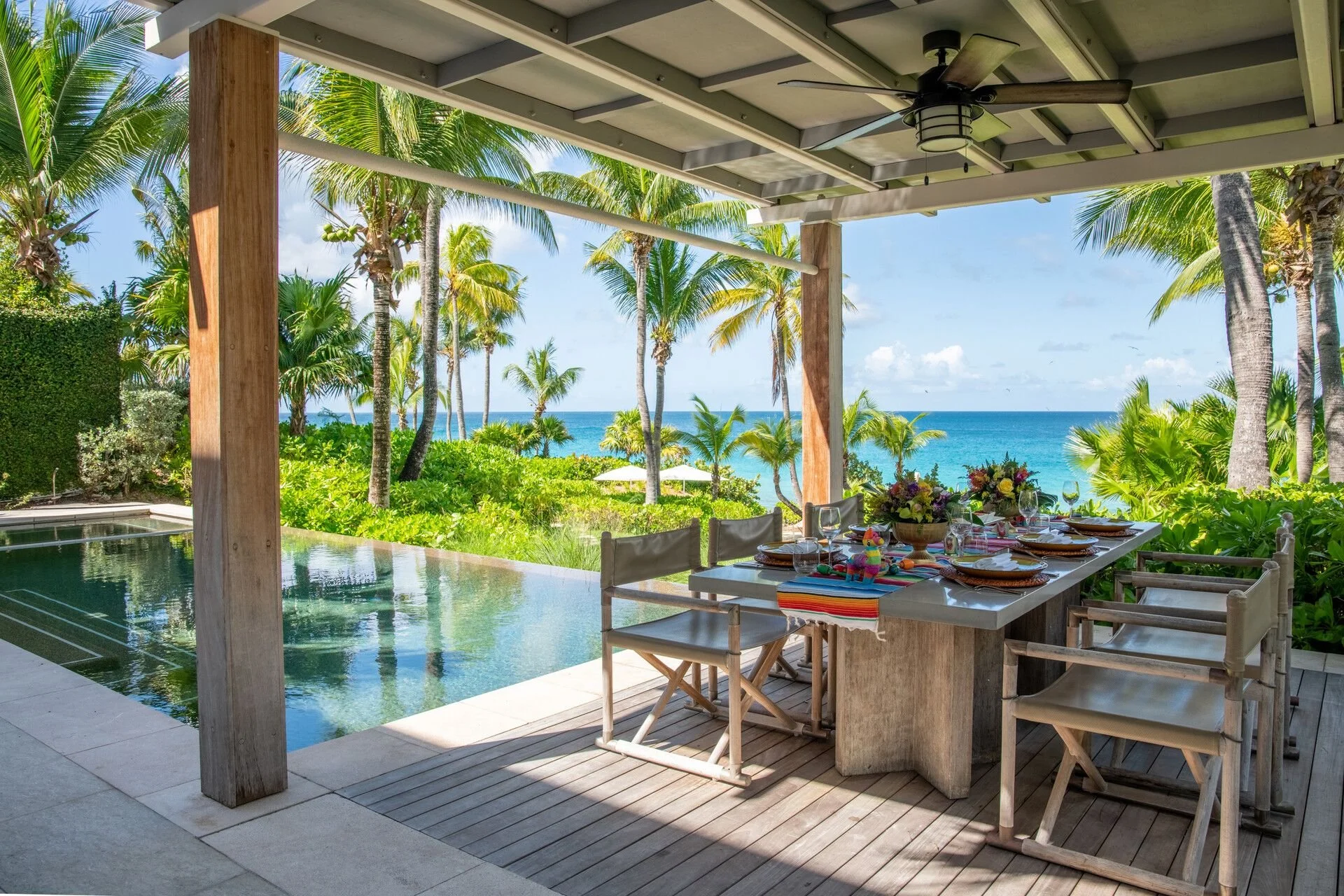 An outdoor dining area on a covered patio with a view of lush tropical trees, palm trees, and the ocean in the background. The dining table is set with plates, glasses, utensils, and a colorful tablecloth, with a centerpiece of flowers. There is a swimming pool visible on the left side.
