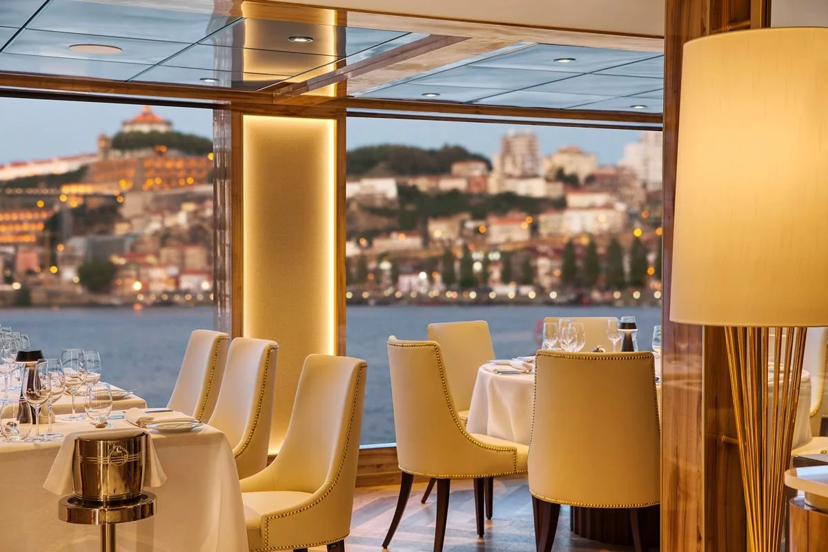Restaurant interior with cream-colored chairs and tables set with glassware, overlooking a river and cityscape at dusk.