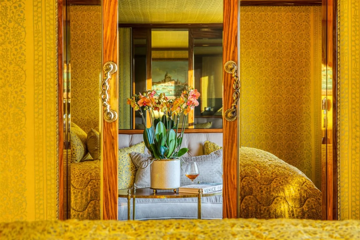 View through a mirror showing a bedroom with yellow-patterned wallpaper, a bed with a yellow bedspread, and a small table with a potted orchid, a glass of rosé wine, and decorative pillows.