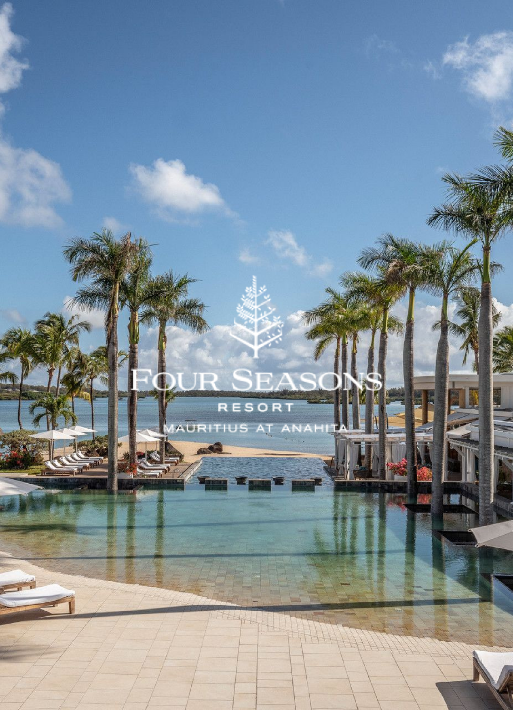 Resort pool area with lounging chairs, tall palm trees, a view of water, and a blue sky with some clouds, at Four Seasons Resort Mauritius at Anahita.