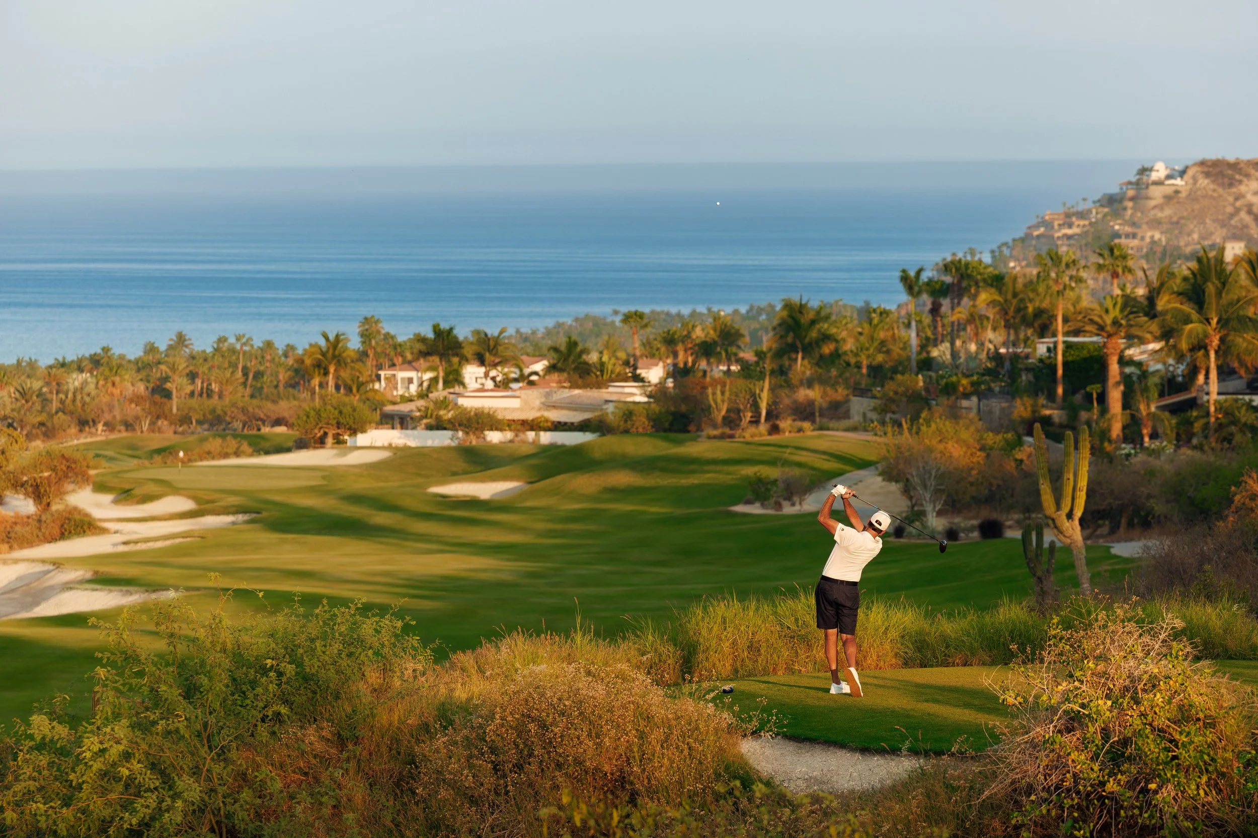 A person playing golf on a lush green golf course by the coast, with houses on a hillside and the ocean in the background.