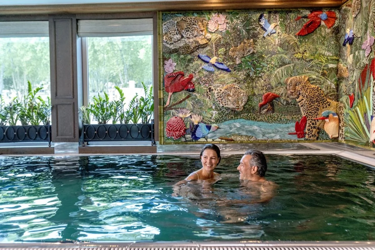 Couple enjoying a swim in an indoor pool with a jungle-themed mural featuring animals and tropical plants on the wall behind them.