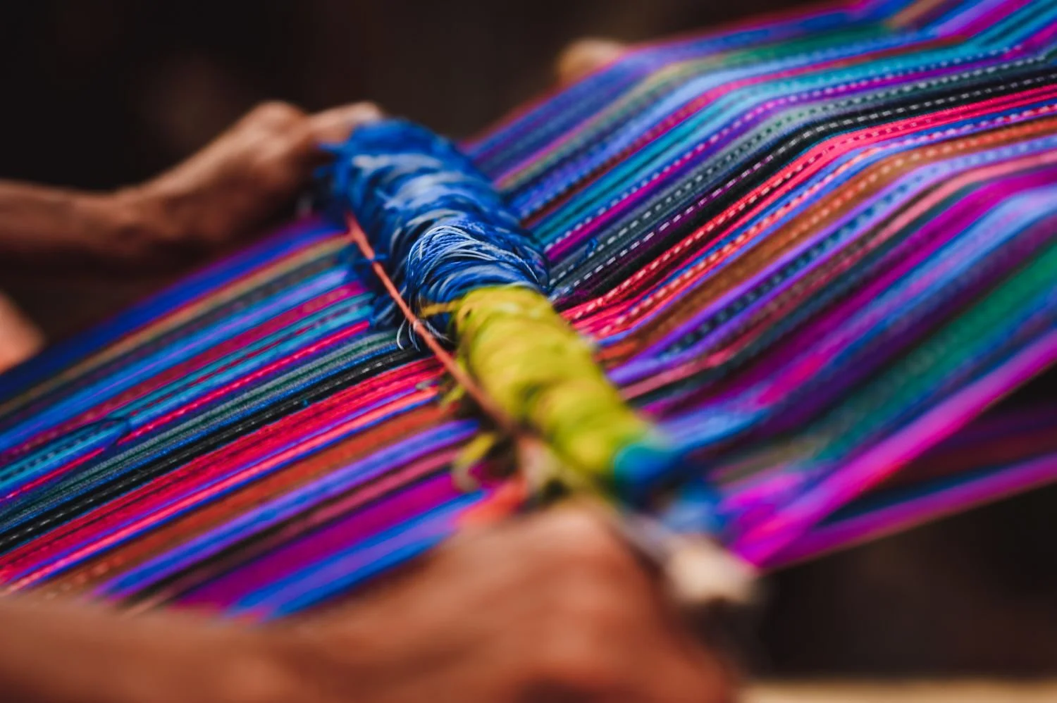 Close-up of vibrant, colorful threads on a weaving loom, with hands of the weaver working on the textile.