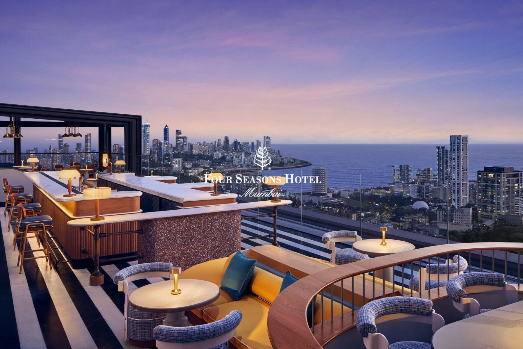 Rooftop lounge at Four Seasons Hotel Mumbai with city skyline and ocean view at sunset, featuring seating areas with lamps, chairs, and tables.