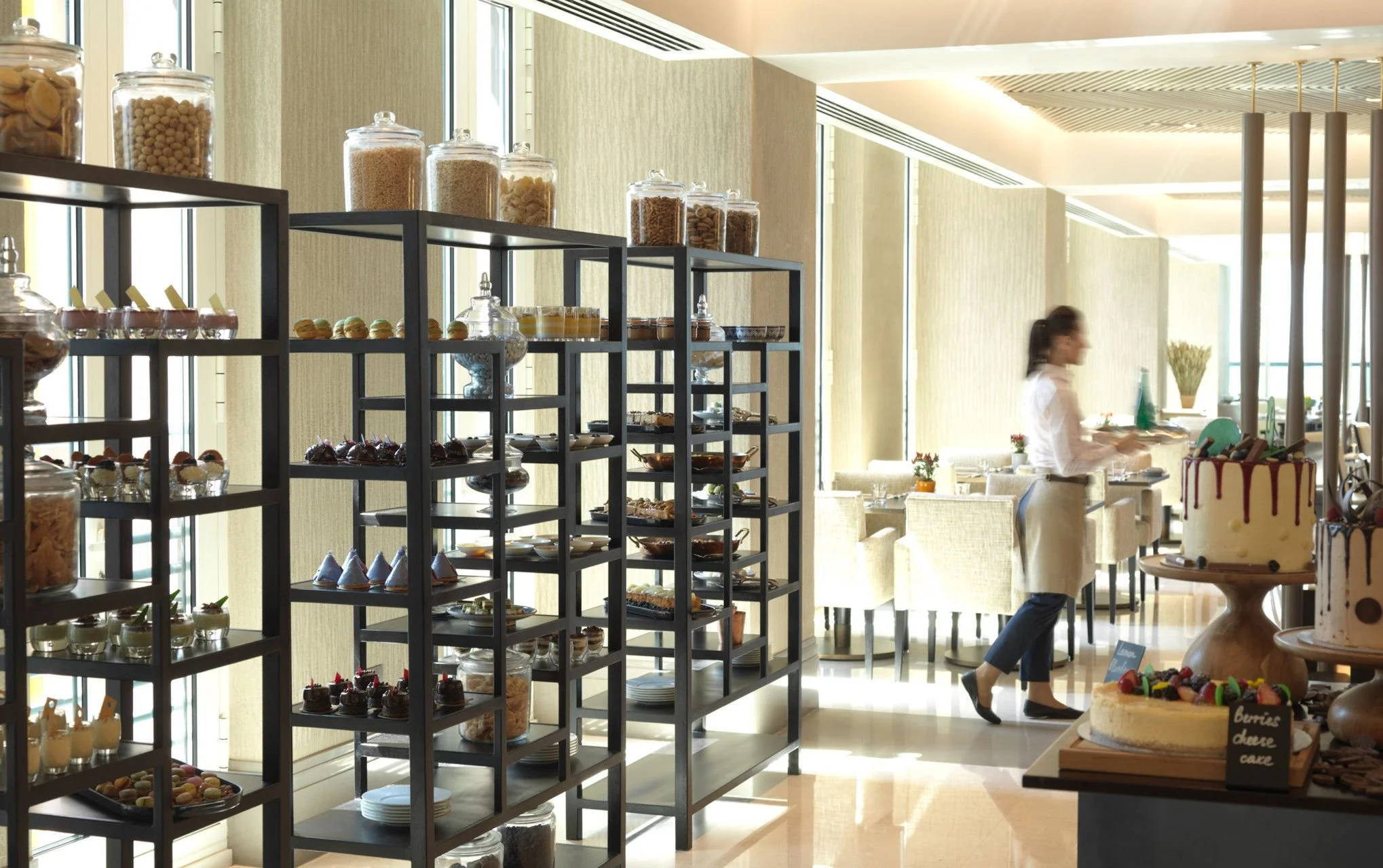 A buffet in a bright dining area with shelves of desserts and baked goods, and a woman serving herself in the background.