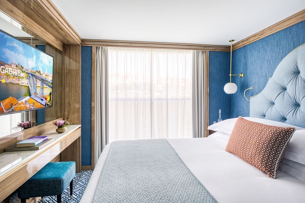 A hotel room with a large bed, blue tufted headboard, and window with sheer curtains. There is a wall-mounted lamp and a glass bottle on a nightstand. A TV is mounted on the wall opposite the bed, and a small desk with books and decorative flowers is nearby.