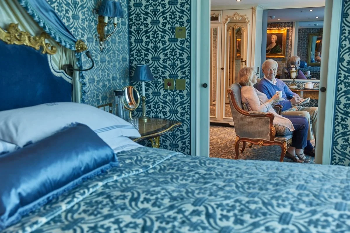 A luxurious bedroom with blue and gold decor facing a sitting area where an elderly couple is sitting, one person holding a cup, with ornate furniture and framed portraits on the walls.