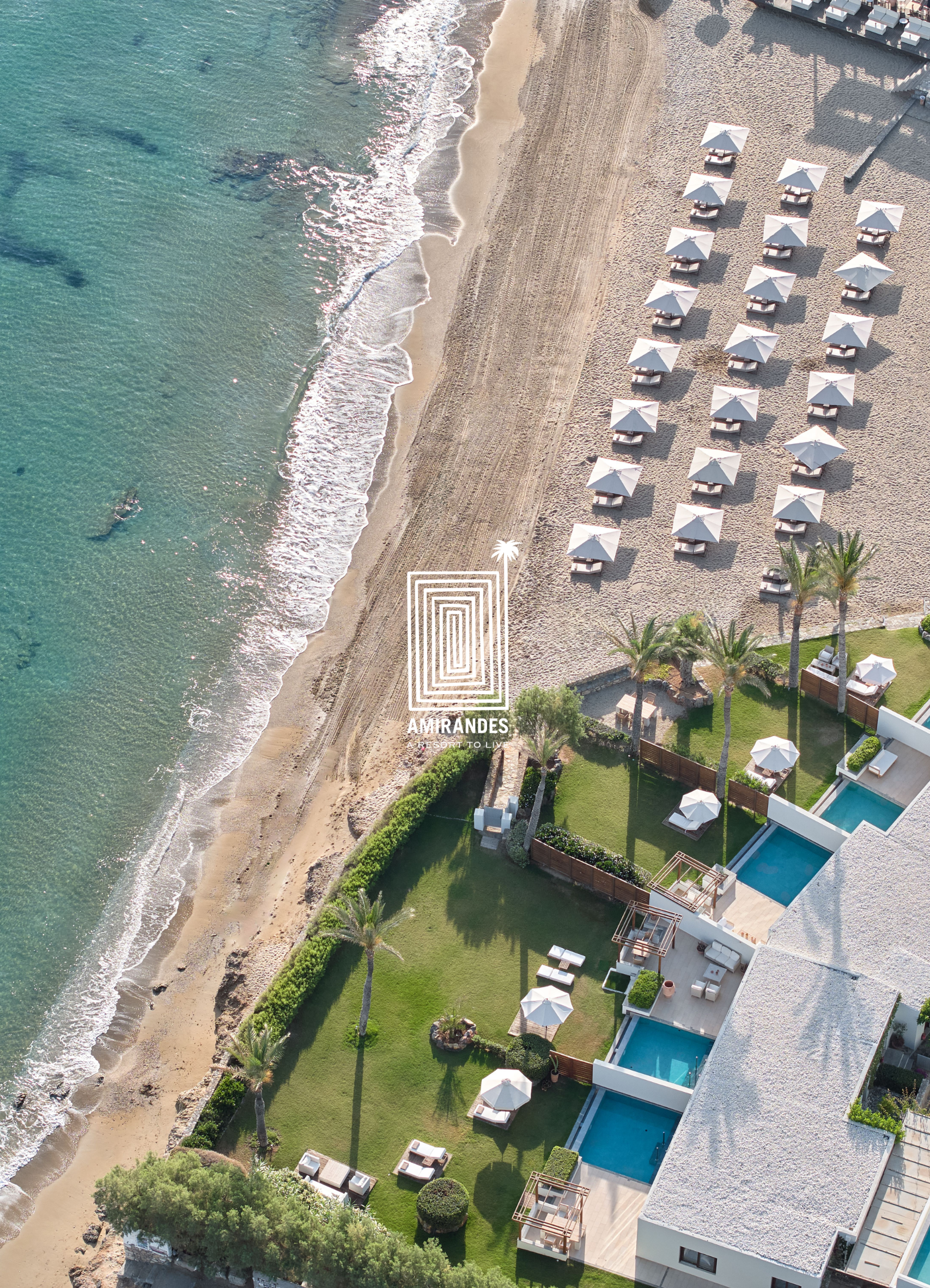 An aerial view of a beachside resort with white tents and umbrellas along a sandy beach and a grassy area with palm trees and pool.