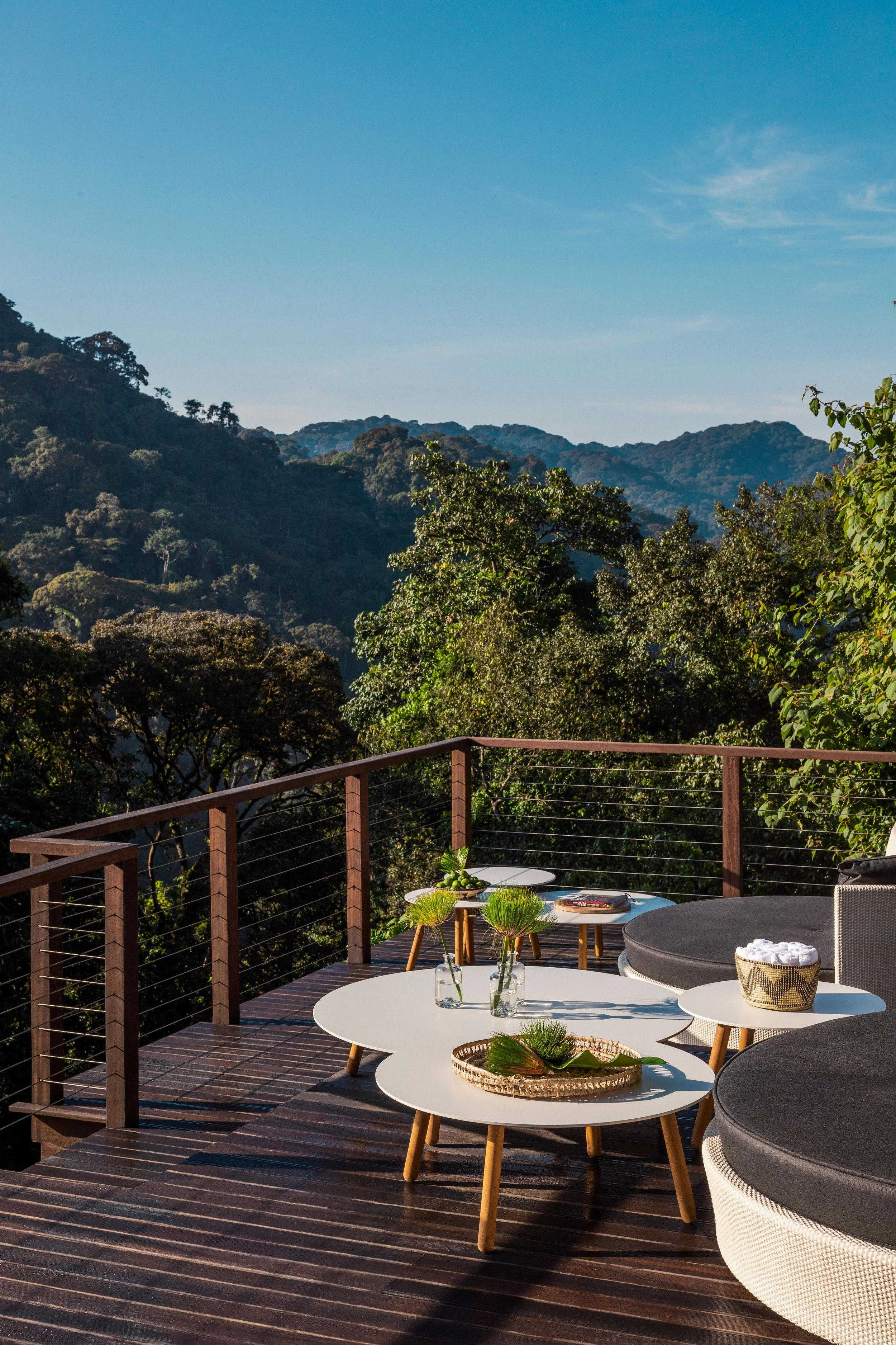 A patio with white and black tables and chairs, surrounded by a wooden railing, overlooking lush green hills and a clear blue sky.