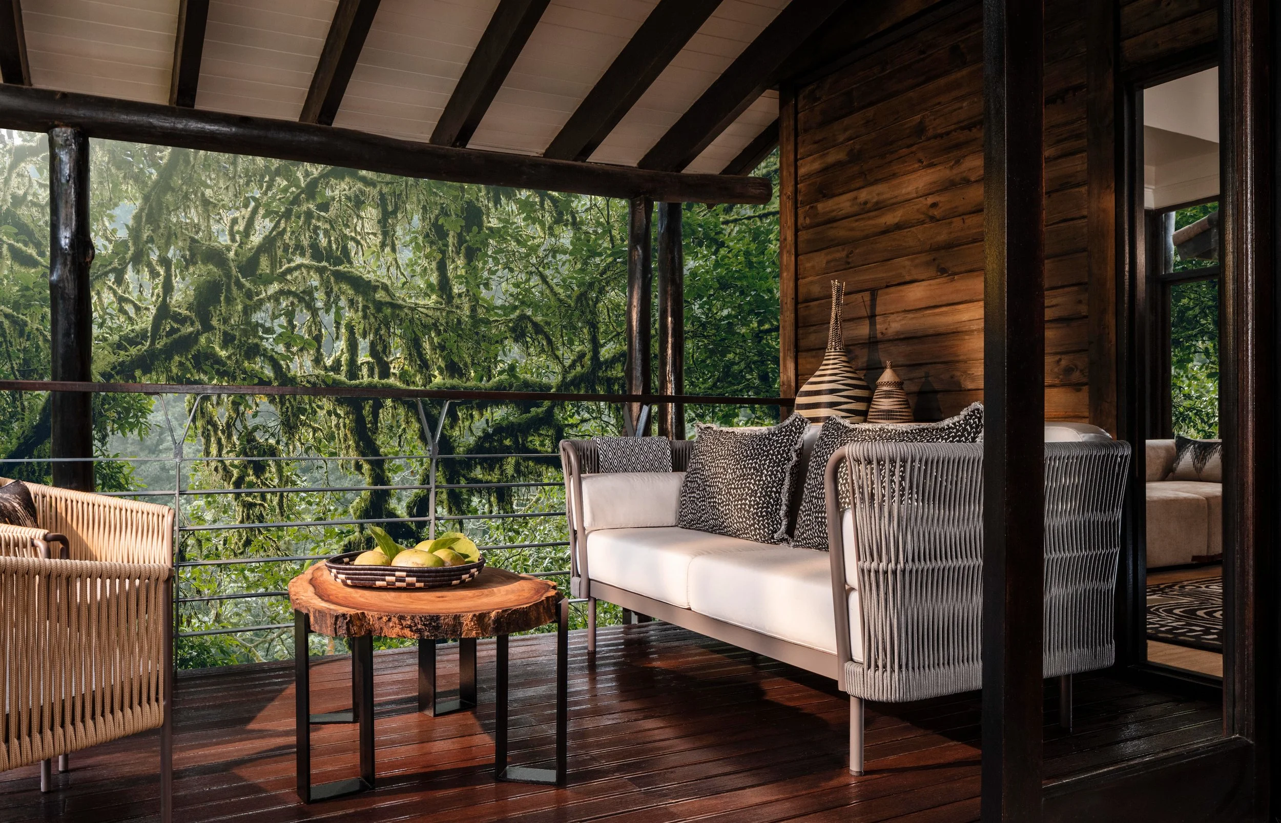 A cozy balcony with wooden walls and ceiling, furnished with white and rattan chairs, a black and white patterned sofa, a round wood table with a basket of fruit, and surrounded by lush green trees visible through the railing.
