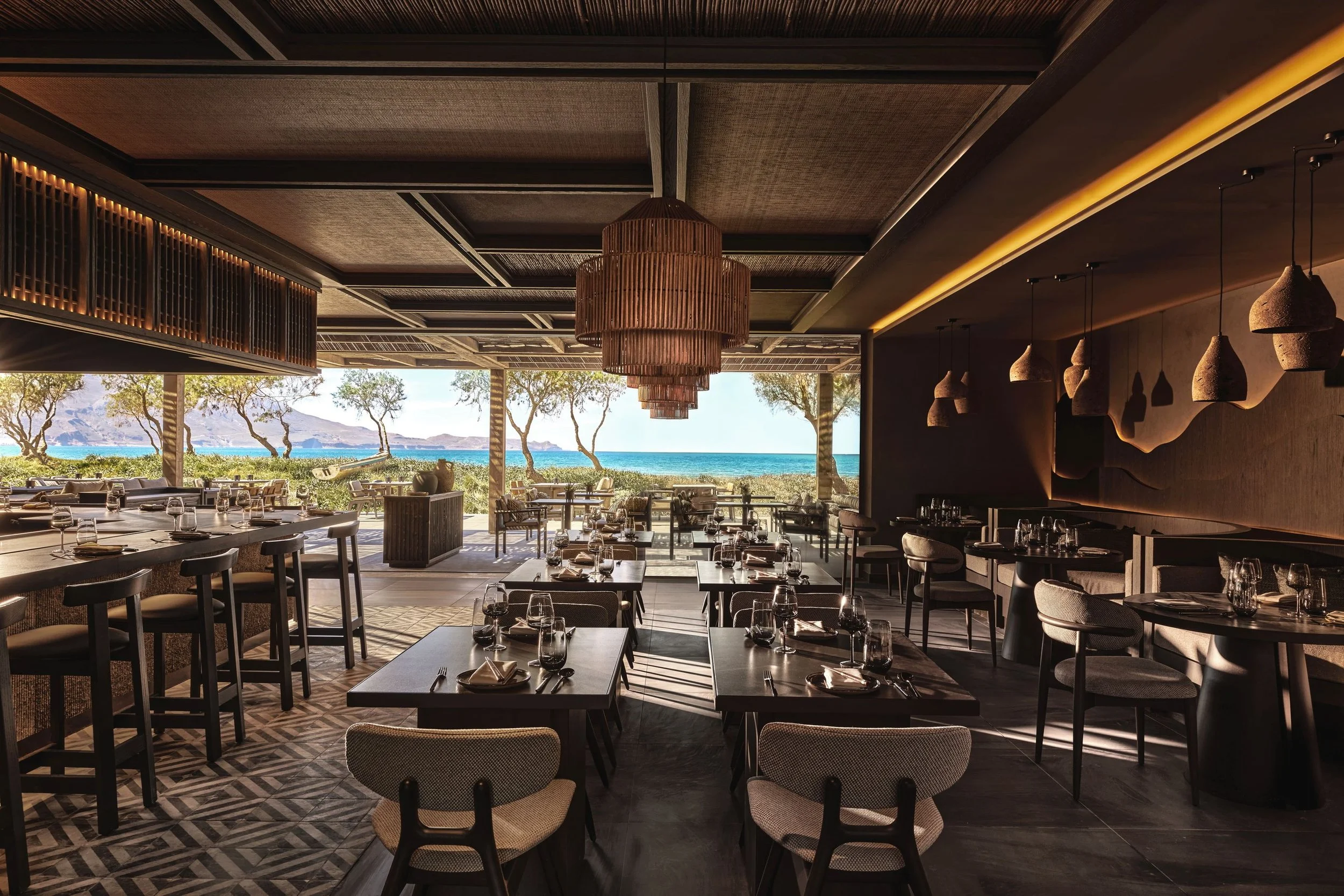 Interior of a modern restaurant with large open windows showing a beach, trees, and mountains in the background. The dining area includes tables set with glassware, napkins, and cutlery, with stylish hanging lamps and a patterned tile floor.