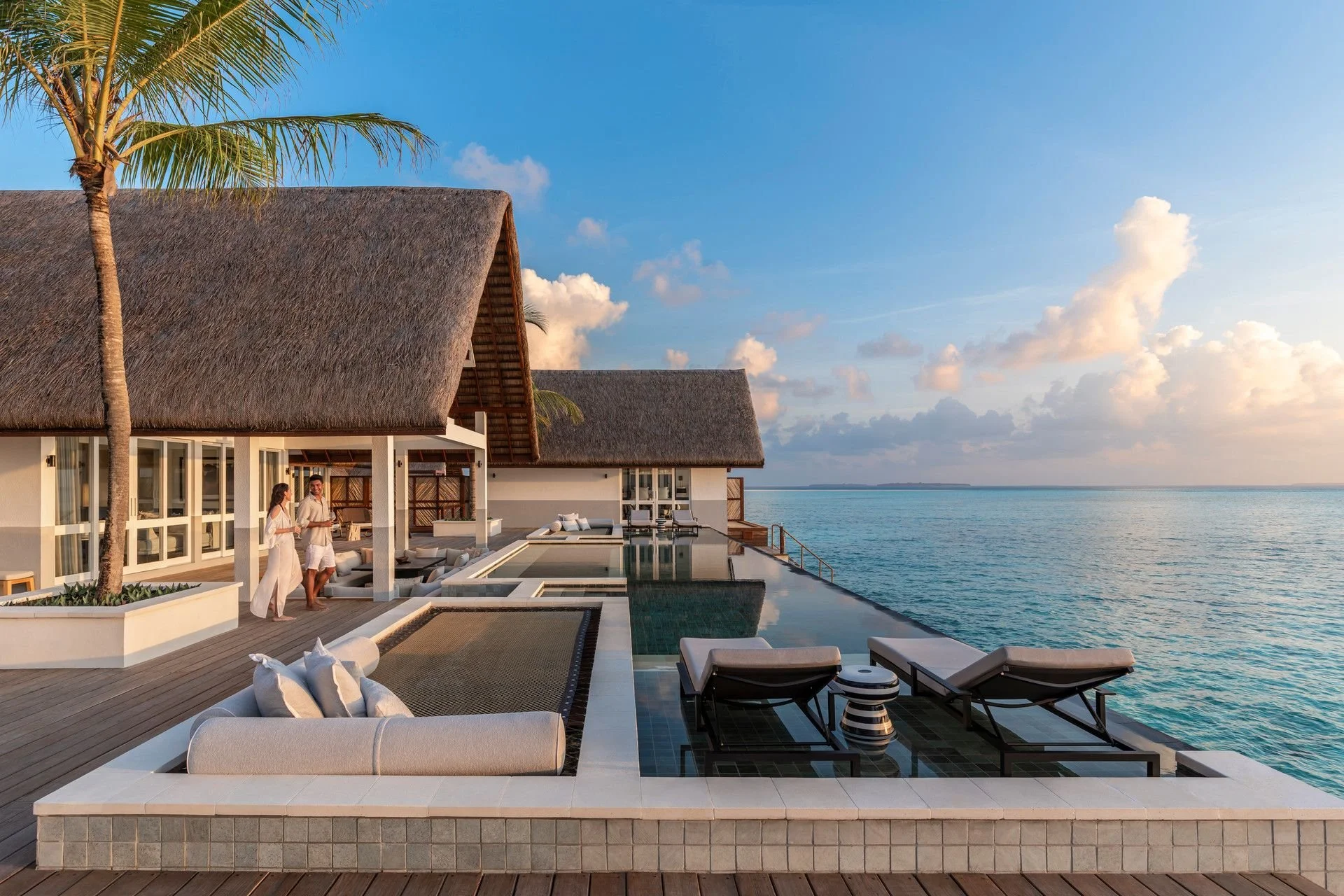 Luxury beachside resort with thatched roofs, outdoor seating, and an infinity pool overlooking the ocean during sunset, with two people standing and talking.