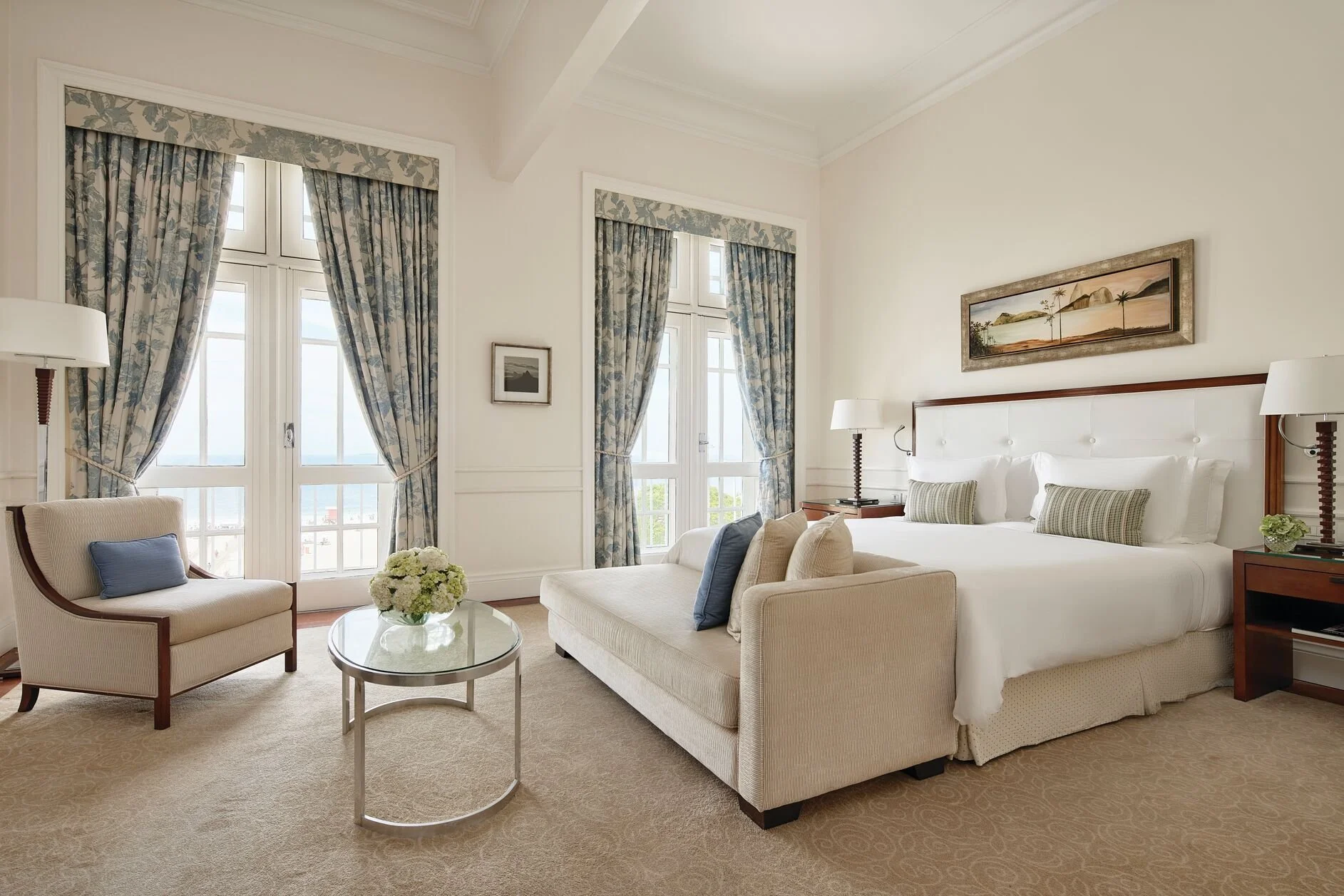 Elegant hotel room with a large bed, nightstands, and table lamps, large windows with floral curtains, sitting area with armchair and small sofa, and wall art of a coastal scene.