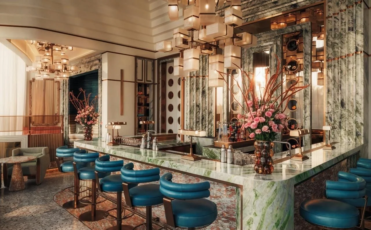 Elegant hotel bar with marble countertops, blue bar stools, decorative lighting, and flower arrangements.