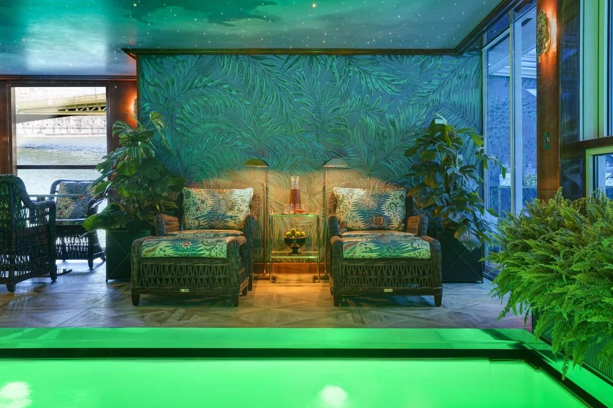 A tropical-themed indoor lounge area with two wicker armchairs with floral cushions, large green plants, and a wall with a leafy pattern mural. There is a small glass table with a lamp and a bowl of lemons between the chairs, and large windows with a view of a deck outside. The ceiling has a starry sky mural, and the room has green accent lighting.