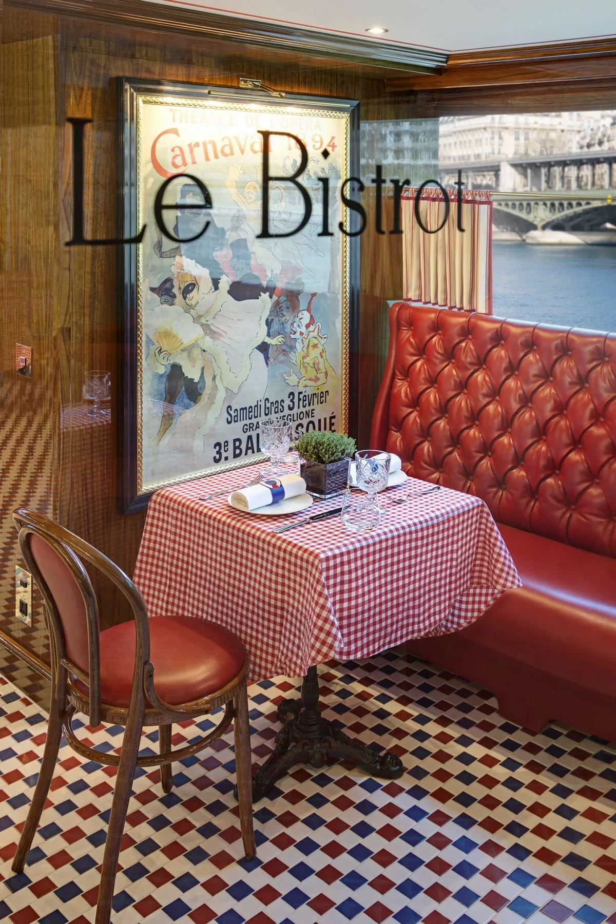 A cozy restaurant booth with a red bench and a small round table covered with a red and white checkered tablecloth, set for a meal with glasses, napkins, and utensils. There is a potted plant on the table and a vintage poster on the window reading 'Le Bistro'. Outside, a river with a bridge and historic buildings are visible.