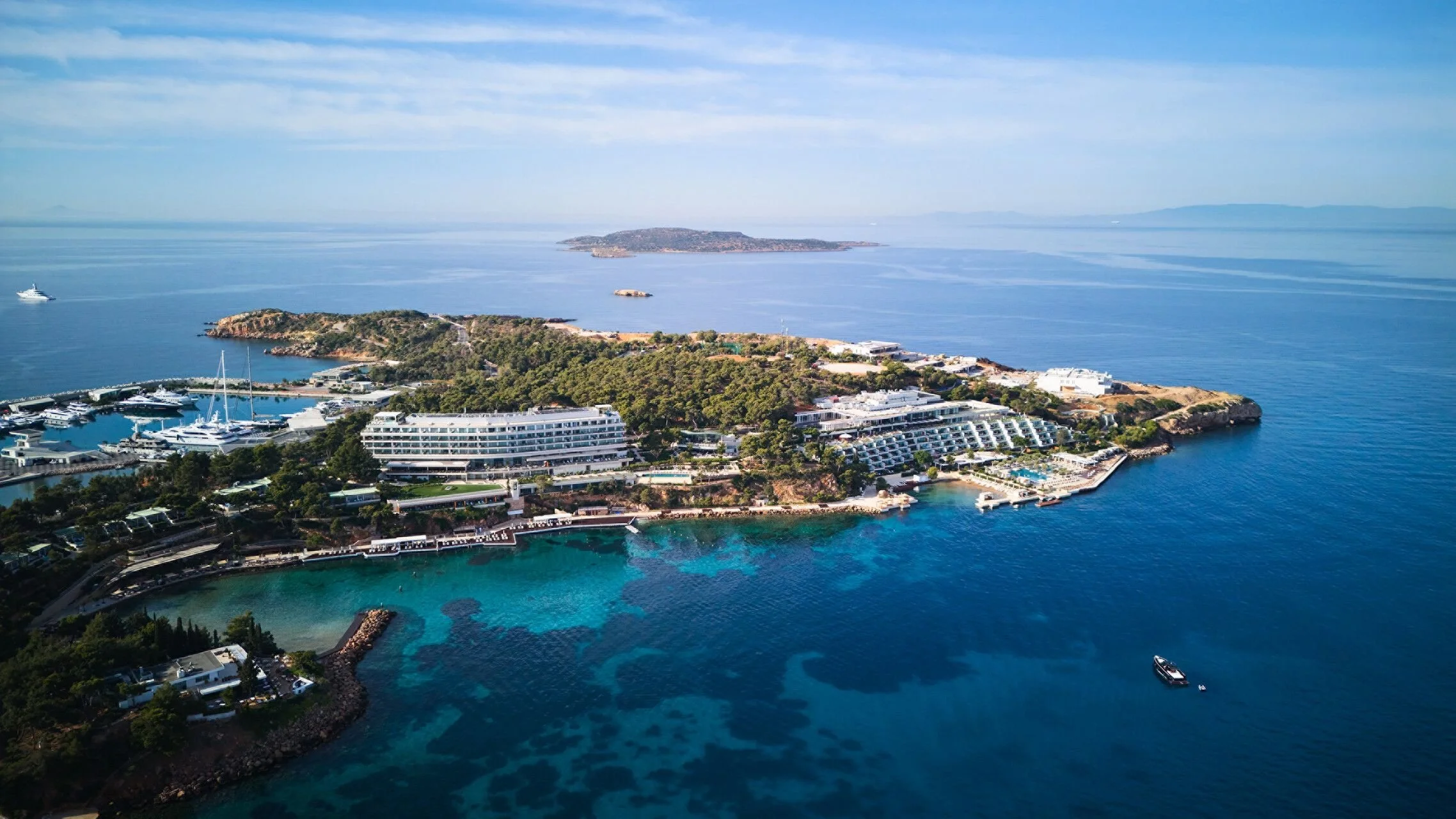 Aerial view of a luxury seaside resort on an island, with white buildings, a marina with yachts, and clear blue water with patches of coral reef.