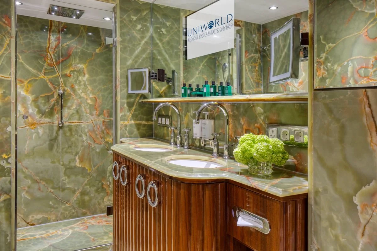 Luxury bathroom with green marble walls and countertop, double sink vanity with wooden base, decorative green flowers, and a glass-enclosed shower.