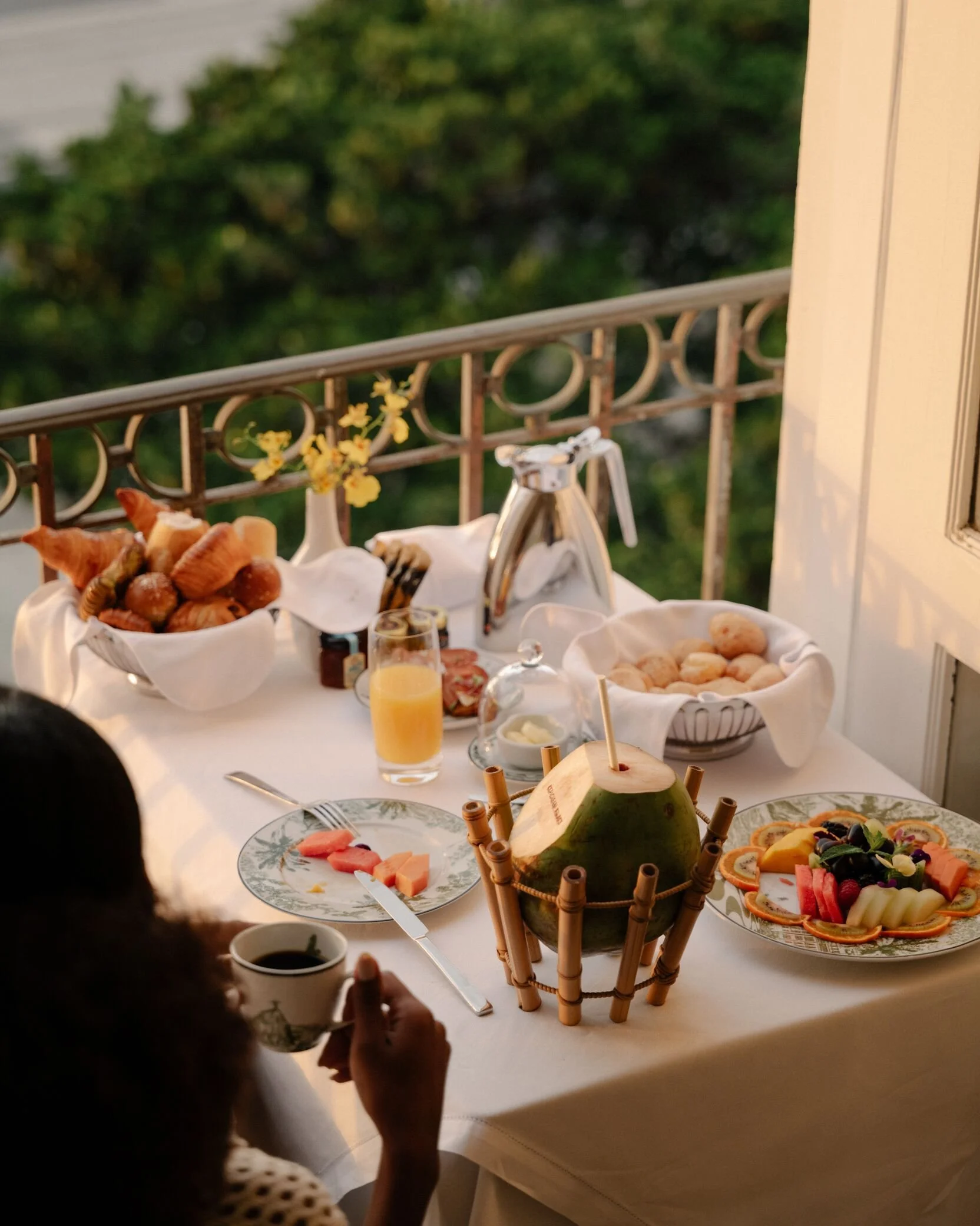 A table set for breakfast on a balcony with a view of greenery. The table has plates of fruit, pastries, a bowl of bread rolls, a glass of orange juice, a coffee cup, and a green coconut with a straw. There are small flowers and a drink dispenser on the table.