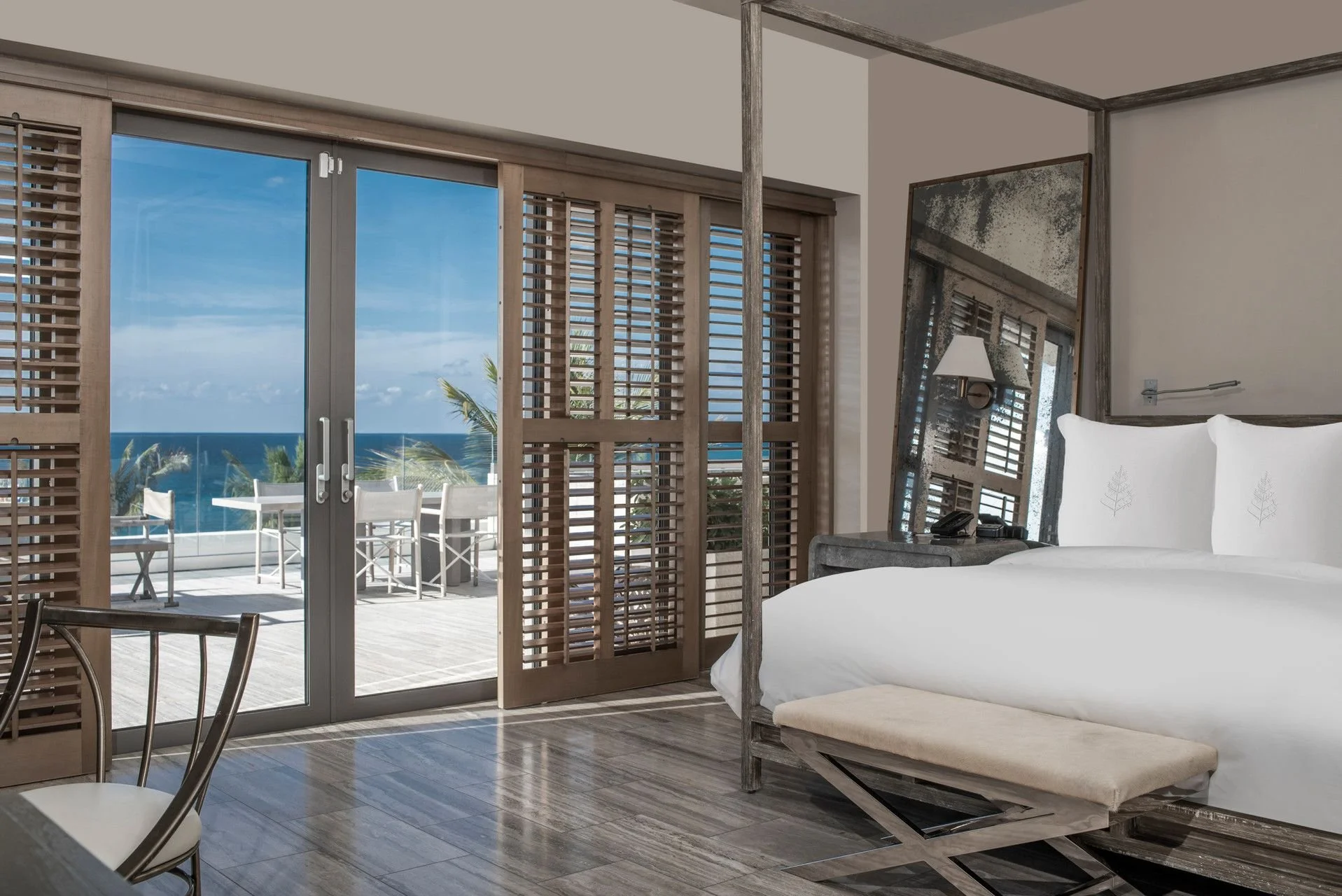 A hotel room with a large bed, a tilted mirror, and a balcony with a view of the ocean and palm trees.