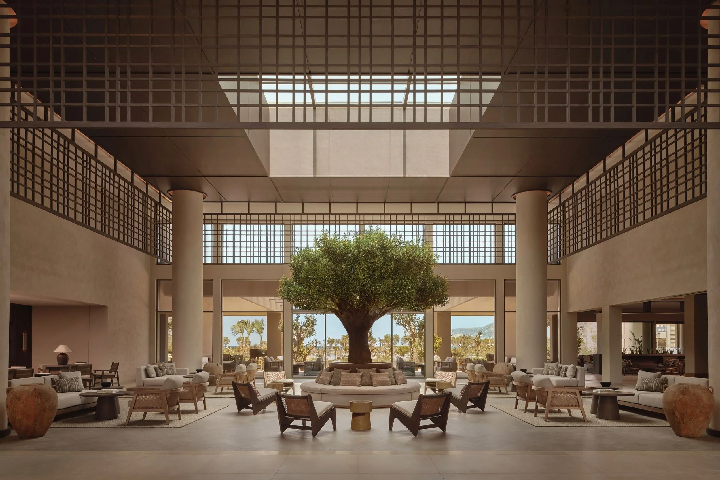 Interior view of a spacious modern lobby with a large central tree, seating area, high ceiling, and grid-like windows allowing natural light.