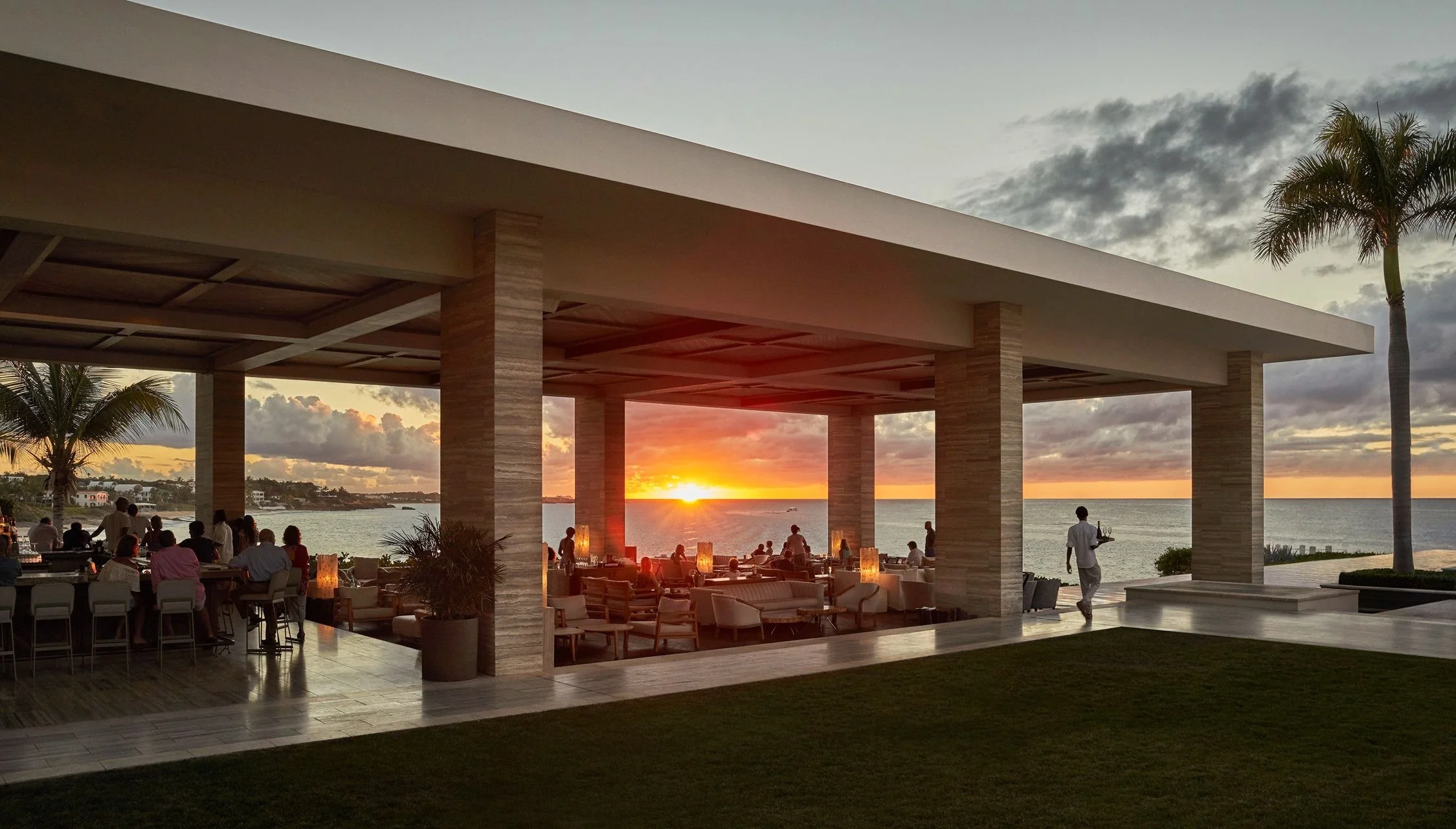 A seaside restaurant with an open-air dining area, guests seated at tables, waitstaff serving, palm trees, and a sunset over the ocean.