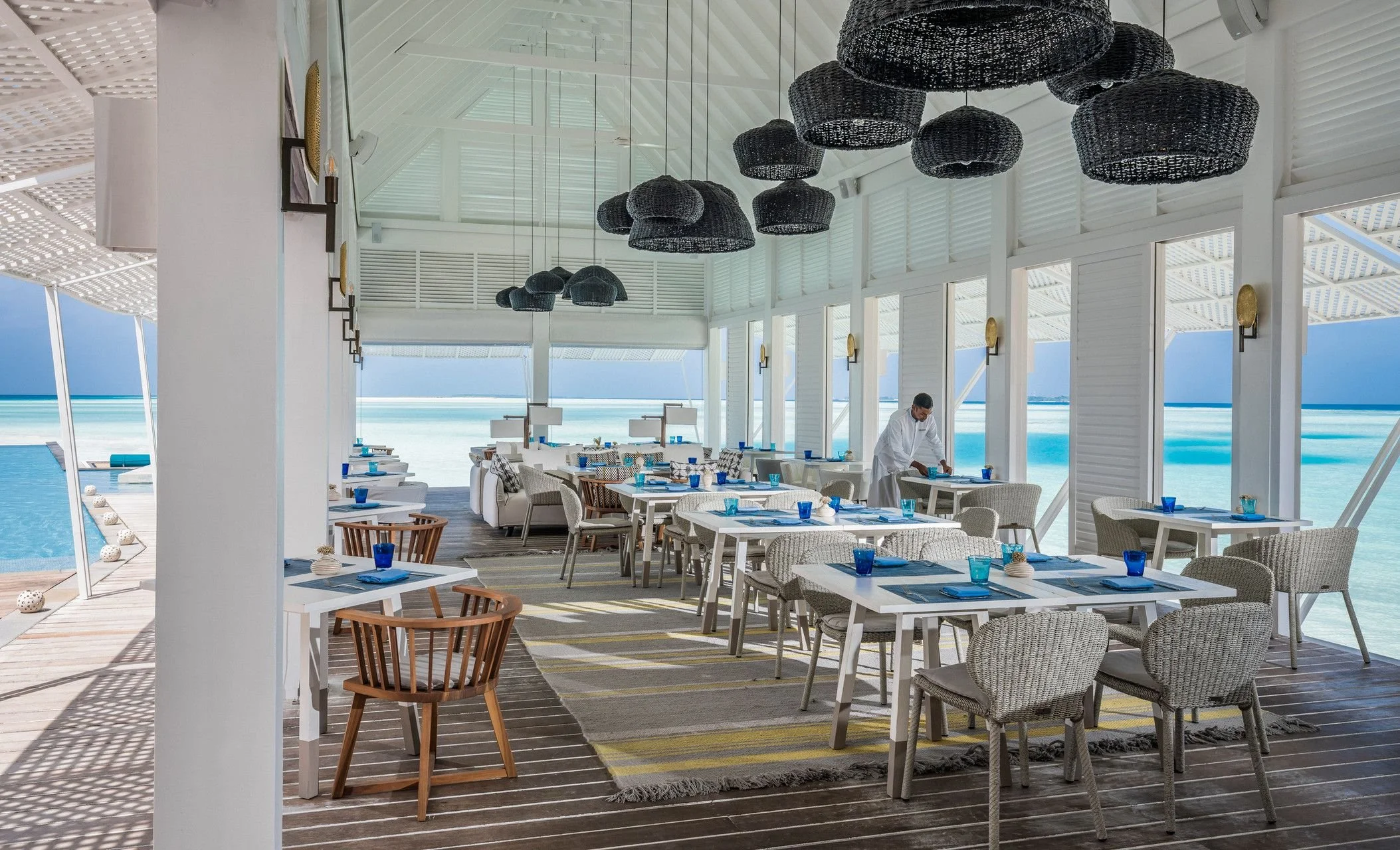 Interior of a beachside restaurant with white walls, black woven pendant lights, and large glass windows showing the ocean. A waiter is setting a table, and the dining area has white and woven chairs with blue glasses and napkins at each place setting.