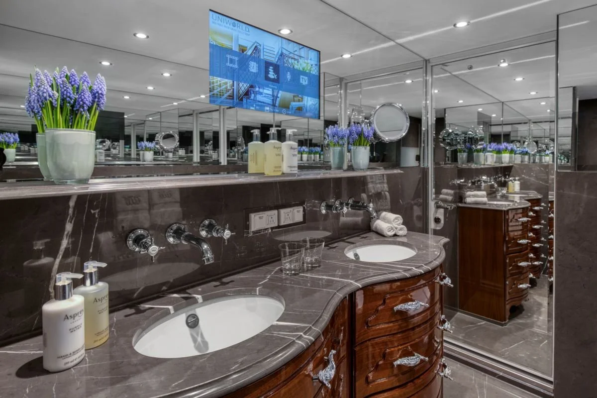 A luxurious bathroom with dual sinks, a large mirror, and a mounted television. The sink area is decorated with multiple vases of purple flowers, bottles of soap, and neatly rolled towels. There are mirrored walls creating a spacious and elegant atmosphere.