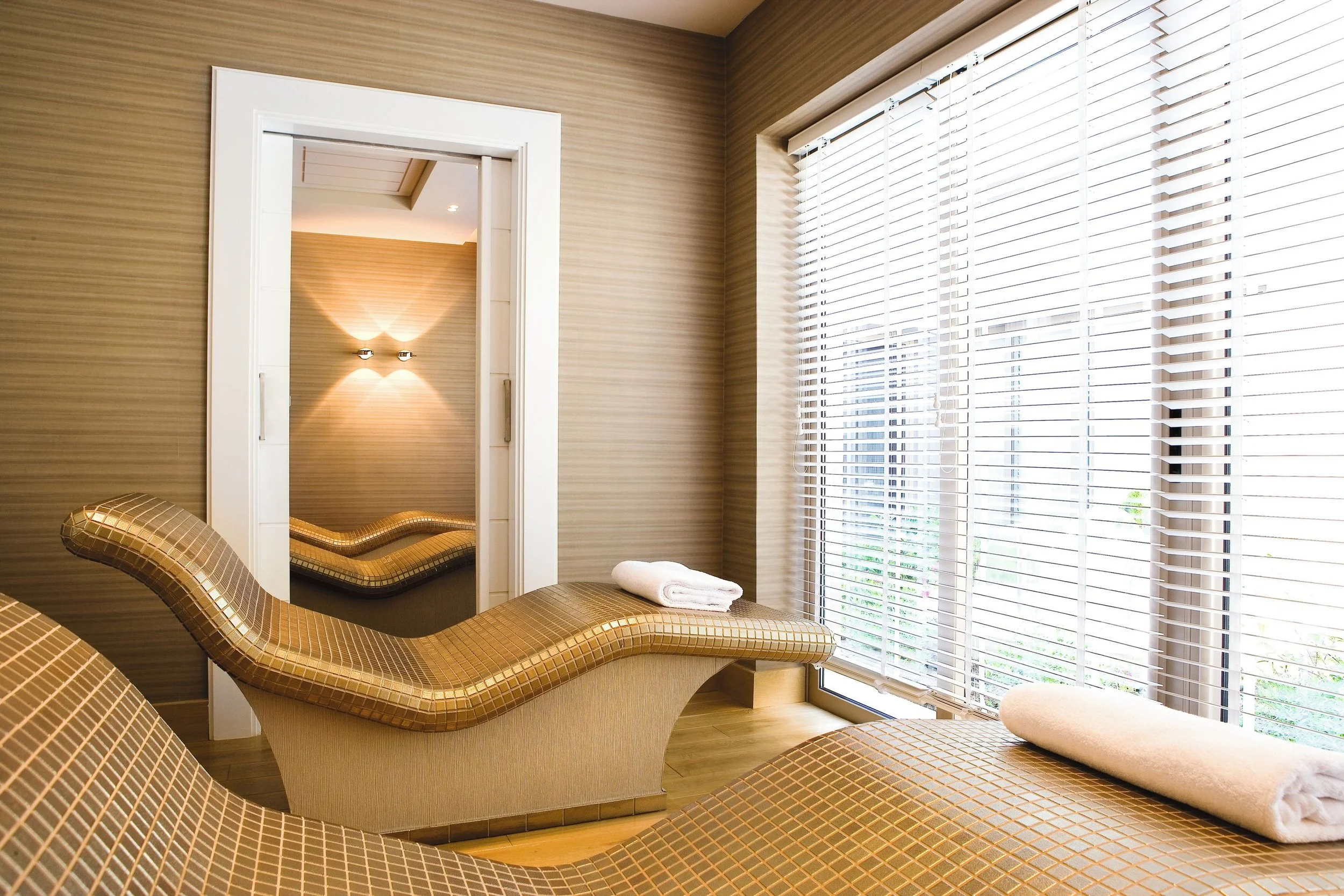Interior of a modern room with beige and brown tones, featuring wooden floors, a large window with white blinds, a curved tan chaise lounge, and a doorway leading to a small room with a wall-mounted light fixture and striped wallpaper.
