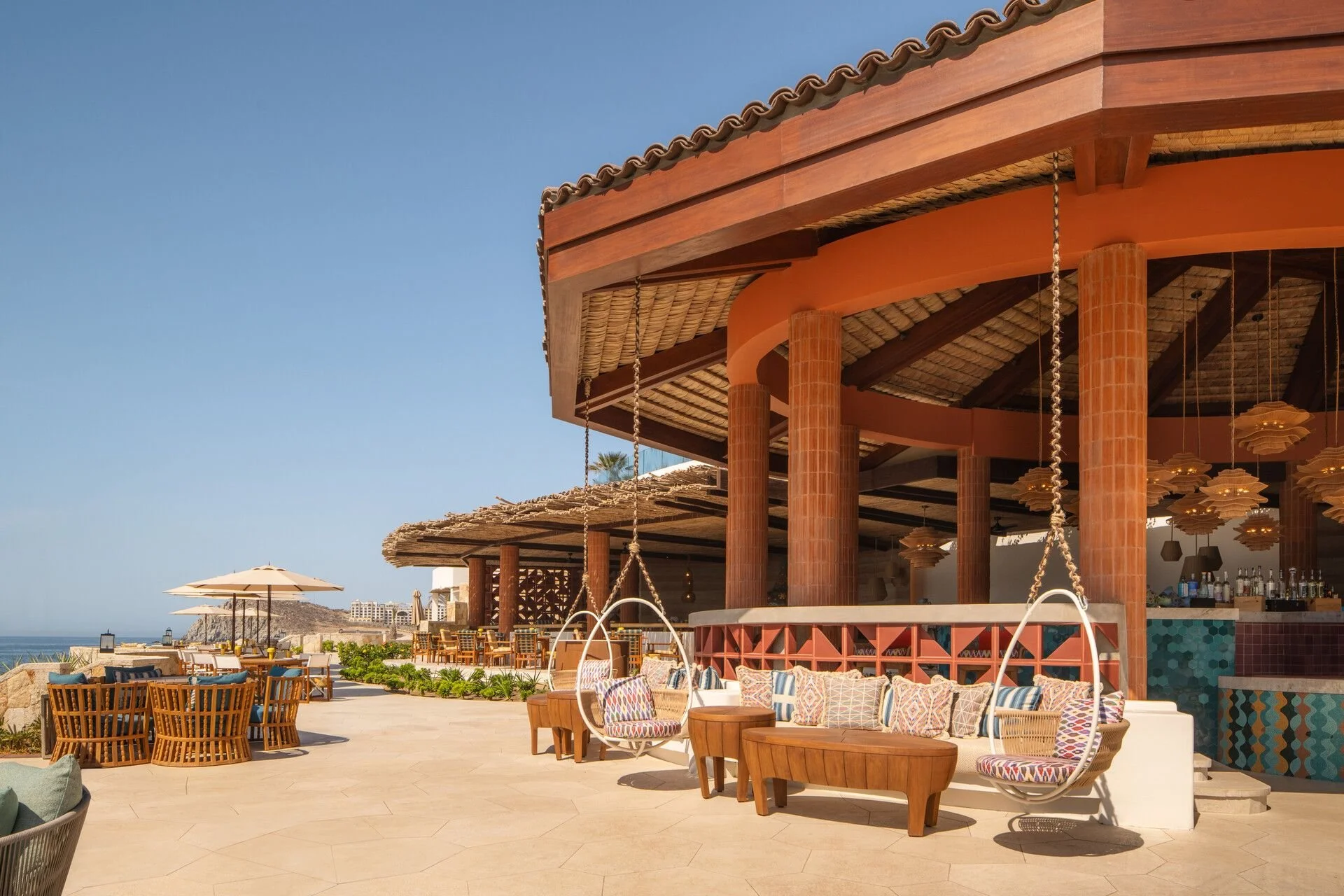 Luxury outdoor bar area with wooden columns, hanging lamps, and colorful seating cushions, overlooking a beach with umbrellas and lounge chairs under a clear blue sky.