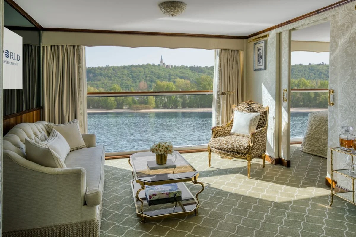 Luxurious hotel room with large windows overlooking a river and forested hills, elegant cream sofa, patterned armchair, coffee table with flowers, and a small bar cart with drinks.