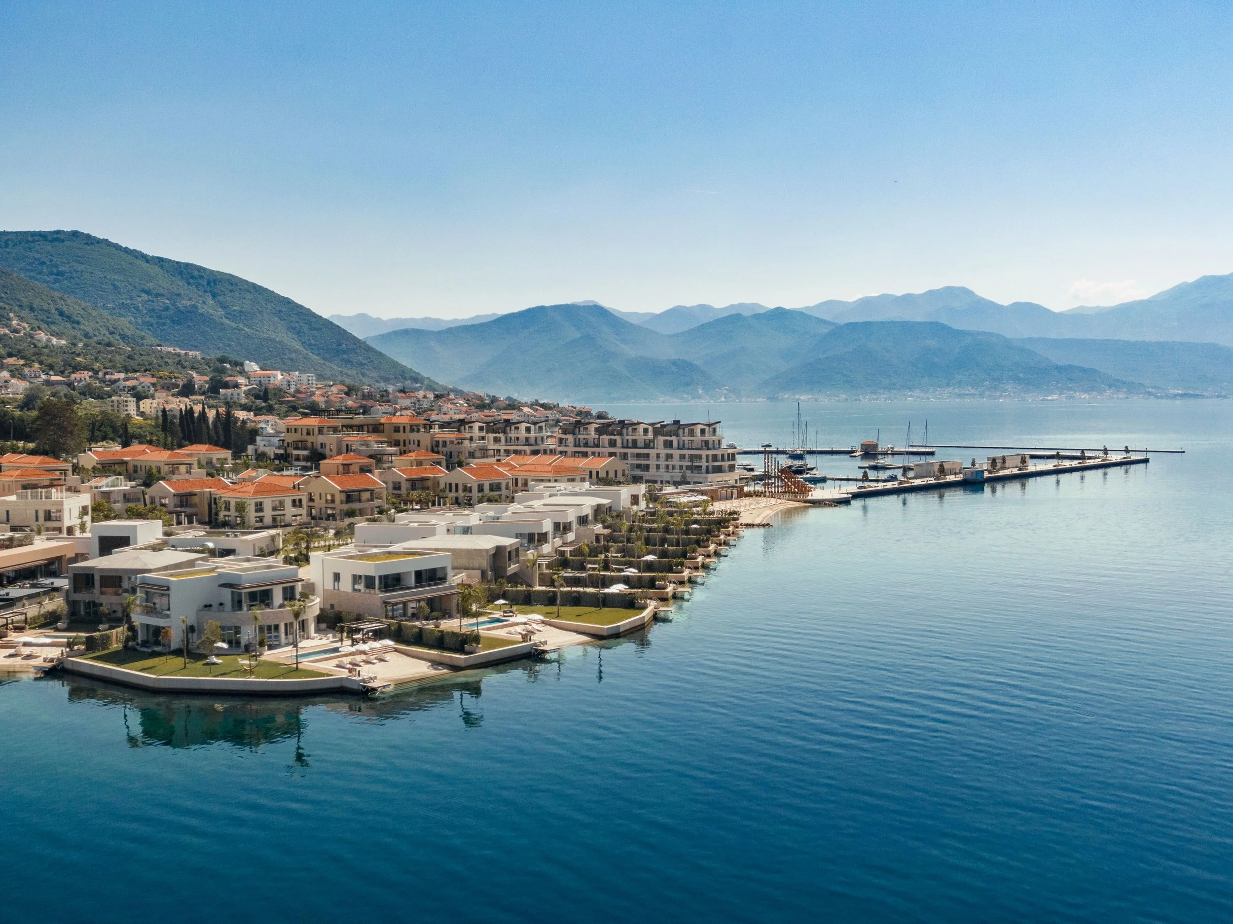 A coastal town with modern houses along the waterfront, surrounded by green hills and mountains in the background. A marina with yachts and boats is visible, with calm water reflecting the clear sky.