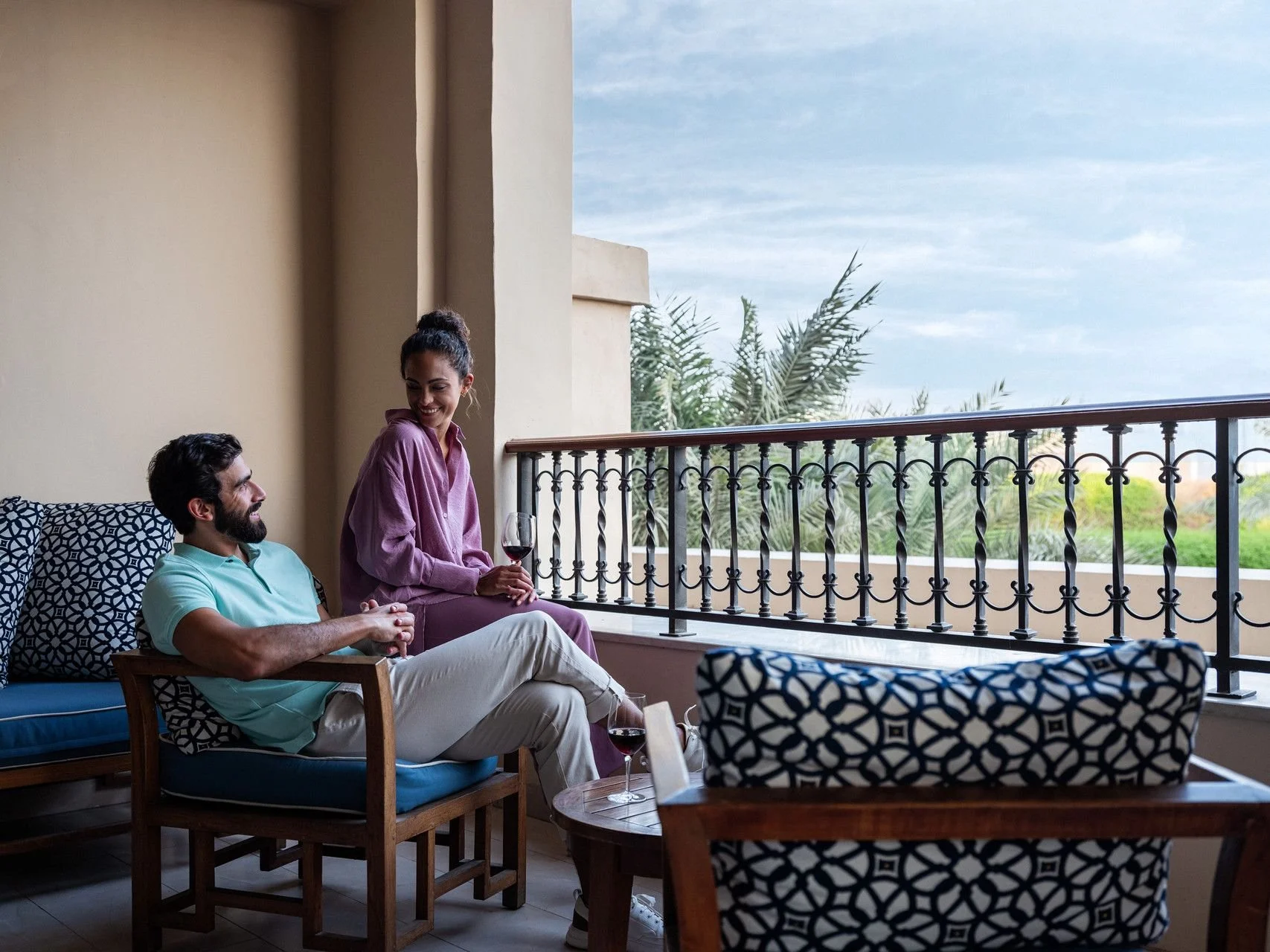 A man and woman relaxing on a balcony, with the woman holding a glass of red wine. They are sitting on comfortable chairs with patterned cushions, and there is a view of palm trees and a partly cloudy sky in the background.