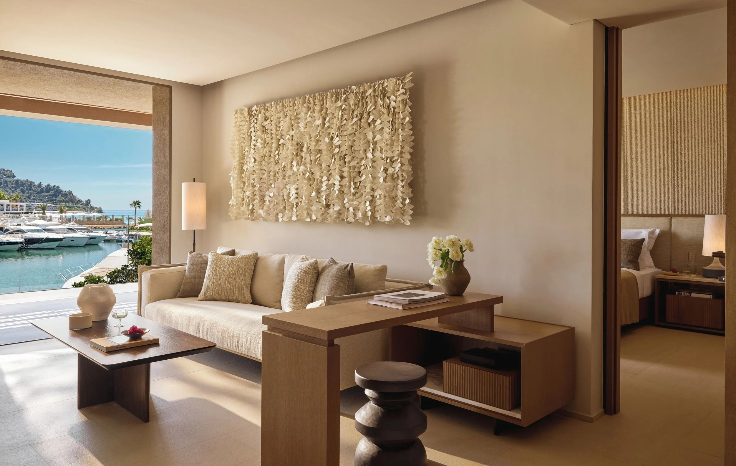 Modern living room with large window overlooking a marina with boats, featuring a beige sofa, abstract wall art, a coffee table, and a bedroom visible through a doorway.
