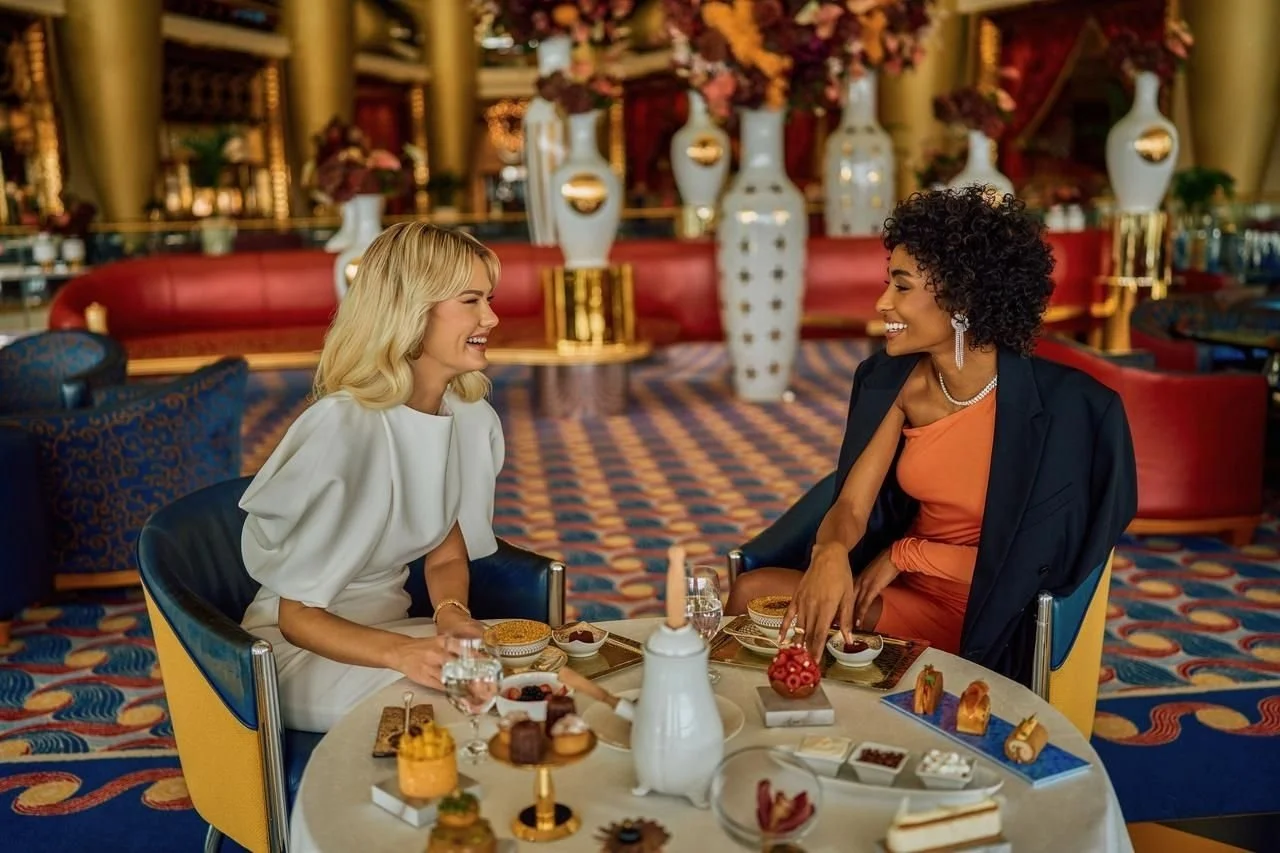 Two women are sitting at a round table in a luxurious restaurant, engaging in conversation and smiling. The table is decorated with various desserts and small dishes, and they are enjoying drinks. The background features elegant vases, large flowers, and ornate decor.