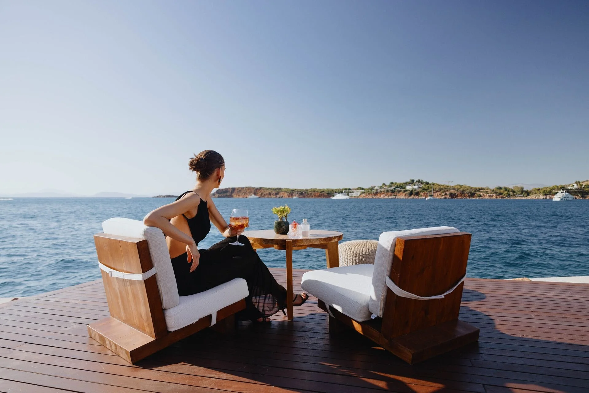 A woman sitting on an outdoor patio overlooking the water, holding a glass of wine, with a small table and table decor nearby on a sunny day.