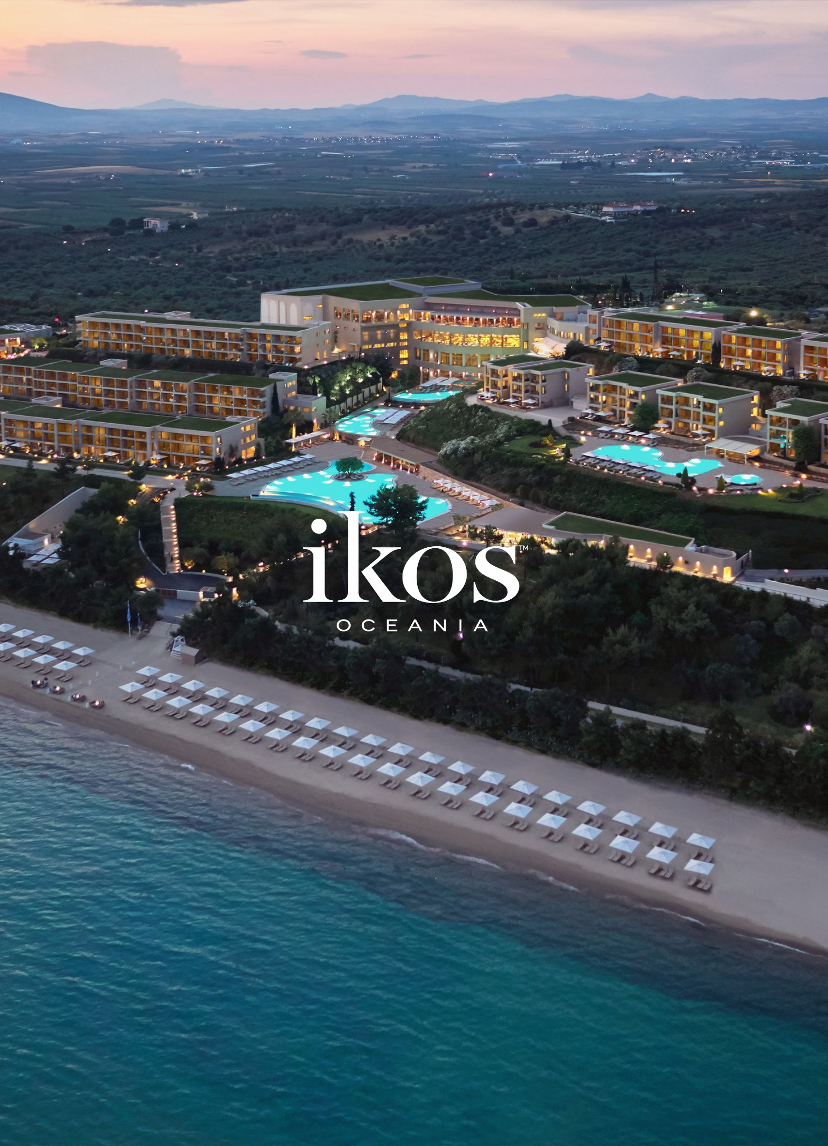 An aerial view of Ikos Oceania resort in Greece at dusk. The resort features multiple beige buildings with green rooftops, swimming pools with blue water, a beach with sun umbrellas, and a coastline with clear turquoise water. The background shows distant mountains and a sunset sky.