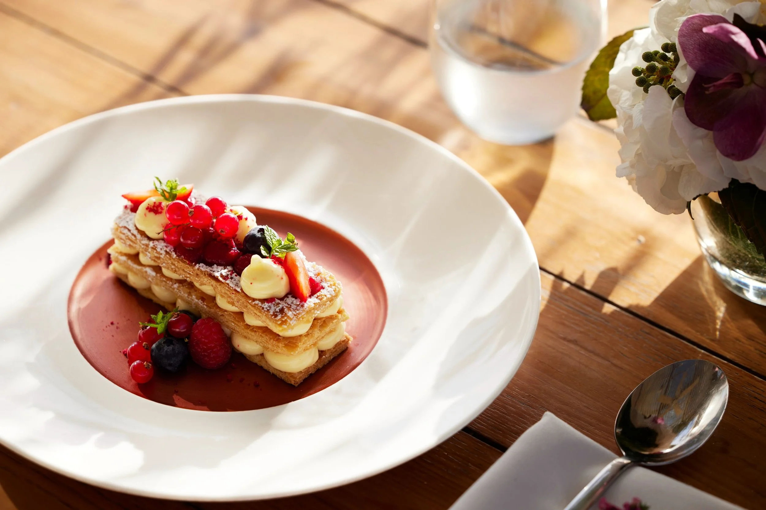 A rectangular layered dessert topped with mixed berries, served on a white plate with a pink sauce underneath, beside a glass of water and a bouquet of flowers on a wooden table.