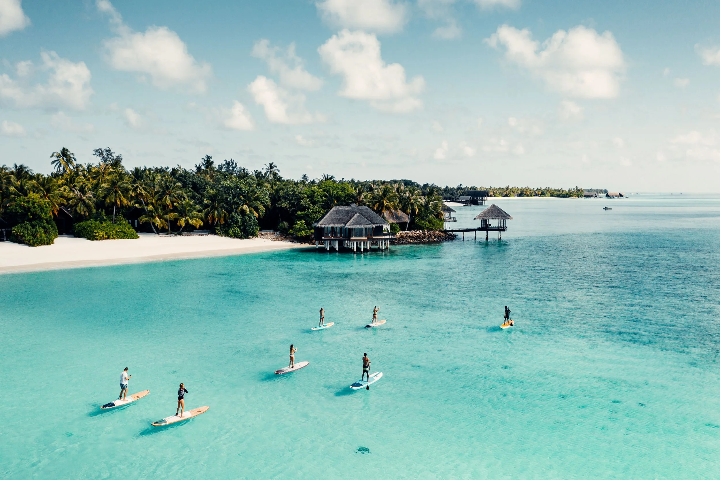 People paddleboarding on turquoise water near a tropical island with palm trees and overwater bungalows.