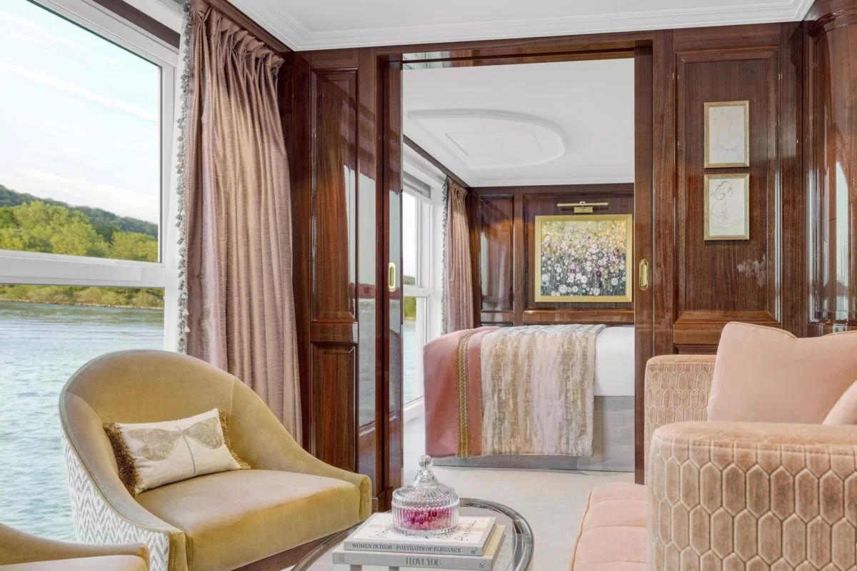 A luxurious living room with large windows overlooking a river and green hills, featuring beige and pink velvet armchairs, a glass table with books and a pink candy dish, and a bedroom with a bed covered in a pink and gold throw.