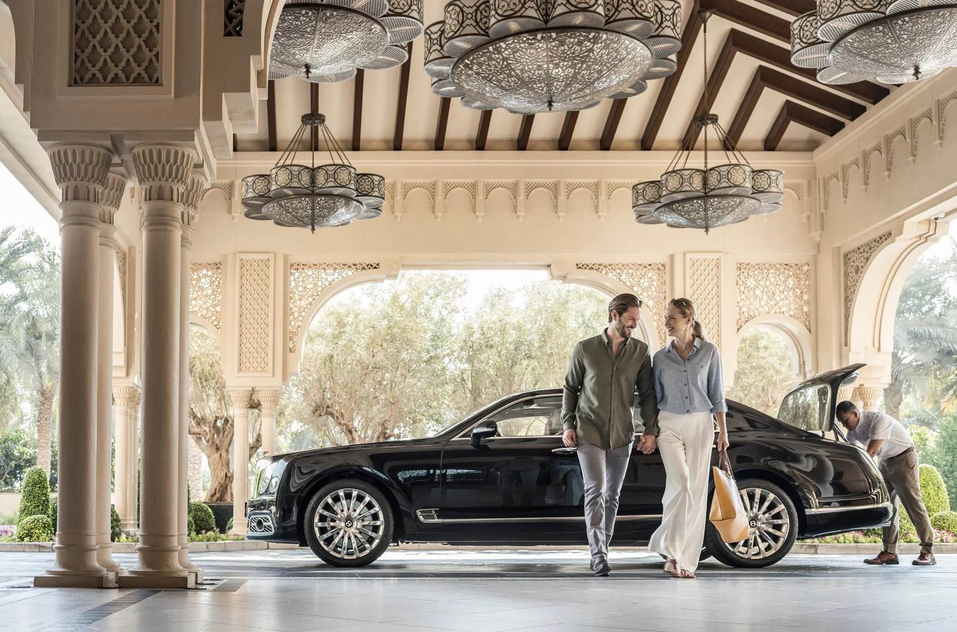 A man and woman are walking hand in hand, smiling, as they leave a luxurious building with a black car parked underneath ornate, decorative ceilings. The man has dark hair and a beard, wearing a gray shirt; the woman has blonde hair tied back, wearing a light blue shirt and white pants. There are other people in the background, and the setting appears elegant, possibly a hotel or upscale residence.