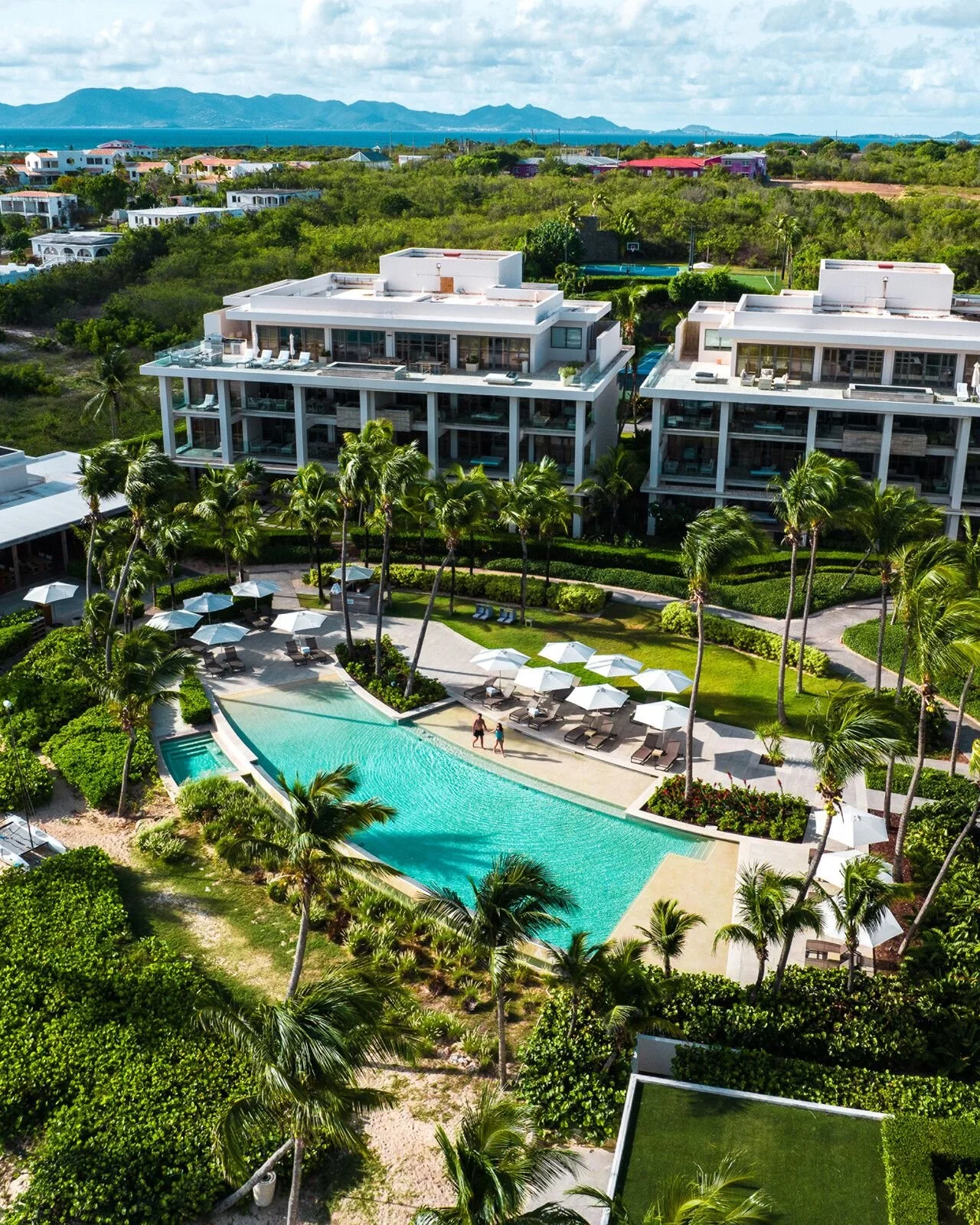 Aerial view of a luxury resort with a swimming pool, palm trees, lounge chairs, modern buildings, and distant mountains and water body.