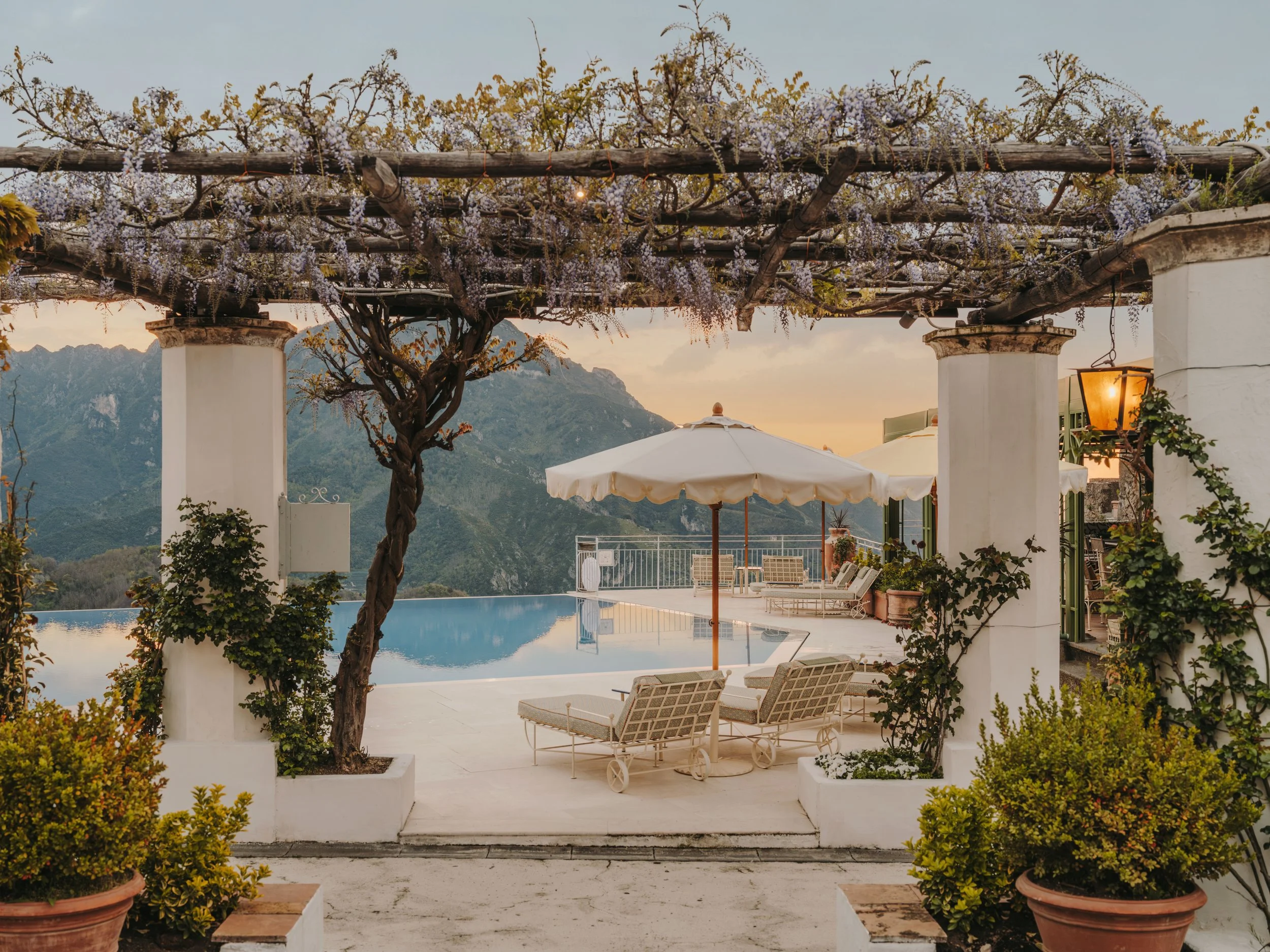 A terrace with white columns, potted plants, a wooden pergola with purple flowering vines, and outdoor furniture including lounge chairs and an umbrella, overlooking mountainous landscape at sunset.