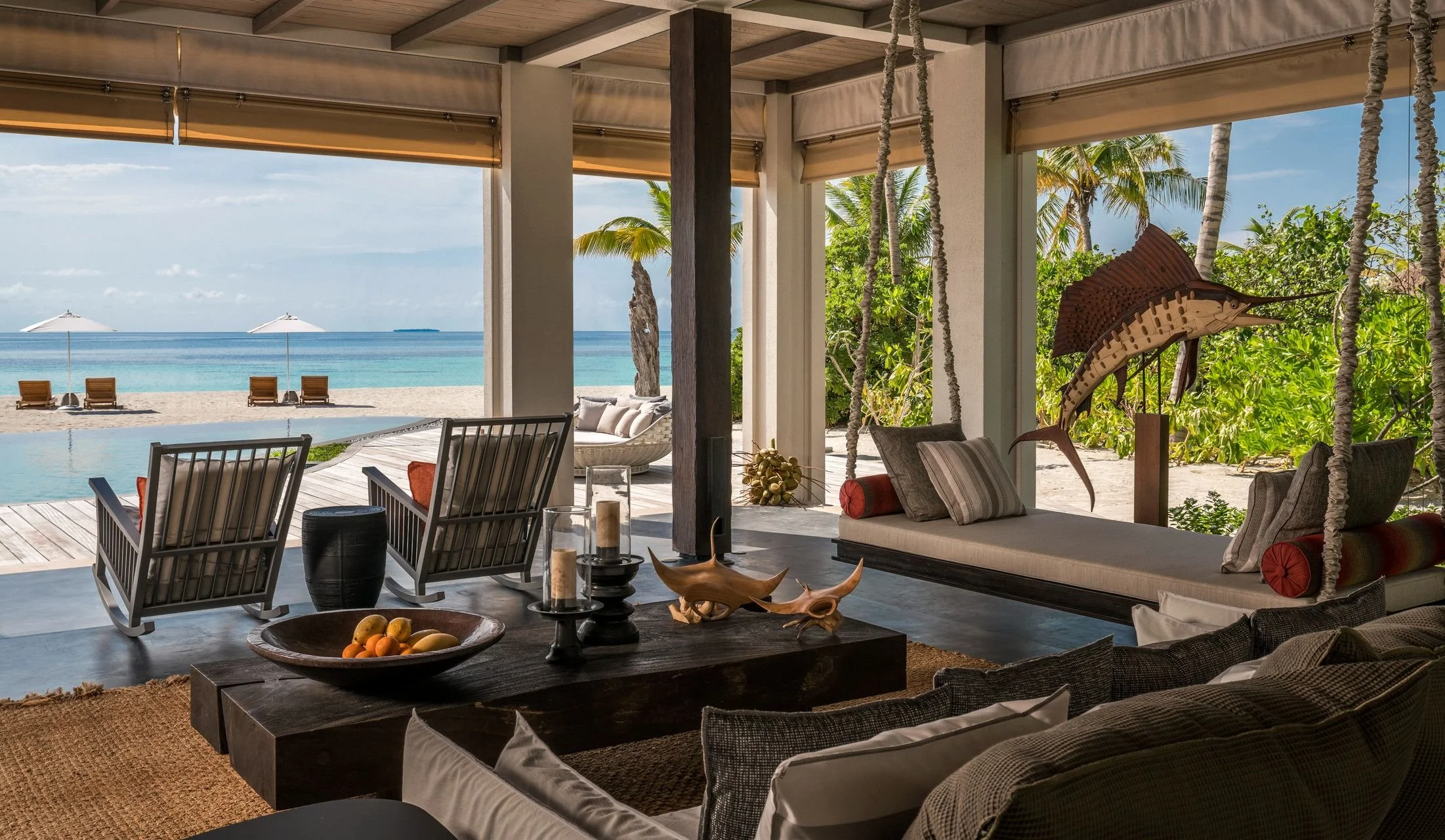 A beachside outdoor lounge area with wooden furniture, pillows, a swing, and tropical plants, overlooking umbrellas and beach chairs on the sand with the ocean in the background.