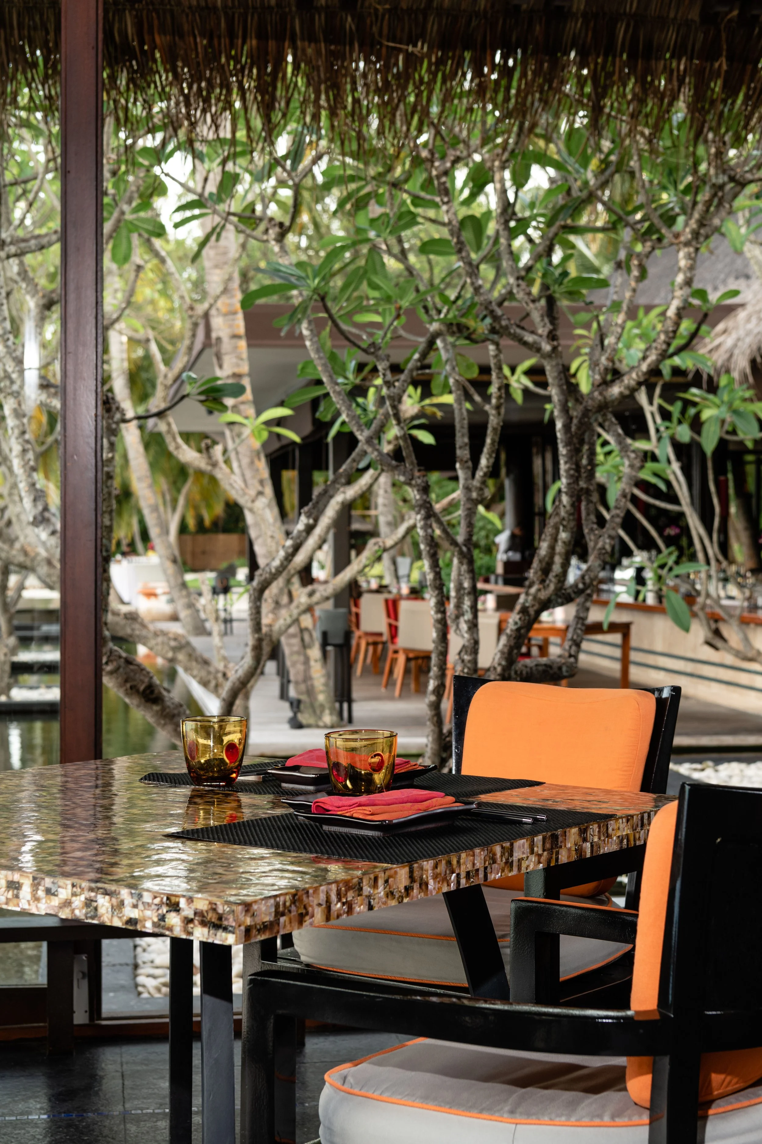 An outdoor restaurant table set for two with orange chairs, black placemats, red napkins, and amber glasses, surrounded by trees and restaurant seating area in the background.