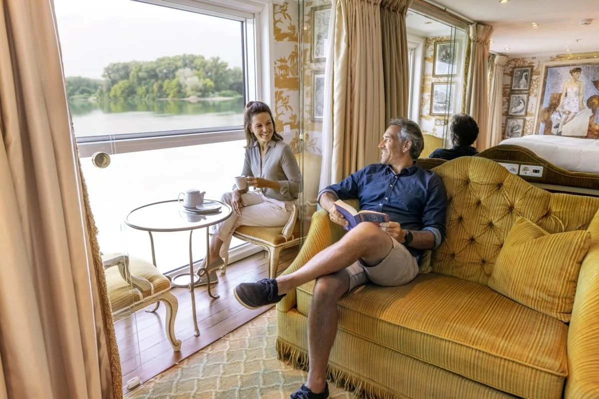 A woman sitting on a chair by a large window with a scenic view of a lake and trees, holding a cup. A man sitting on a yellow upholstered sofa, reading a book, smiling and looking towards the woman. Another person is sitting on a bed in the background of a luxurious, decorated room with framed art and a patterned wallpaper.